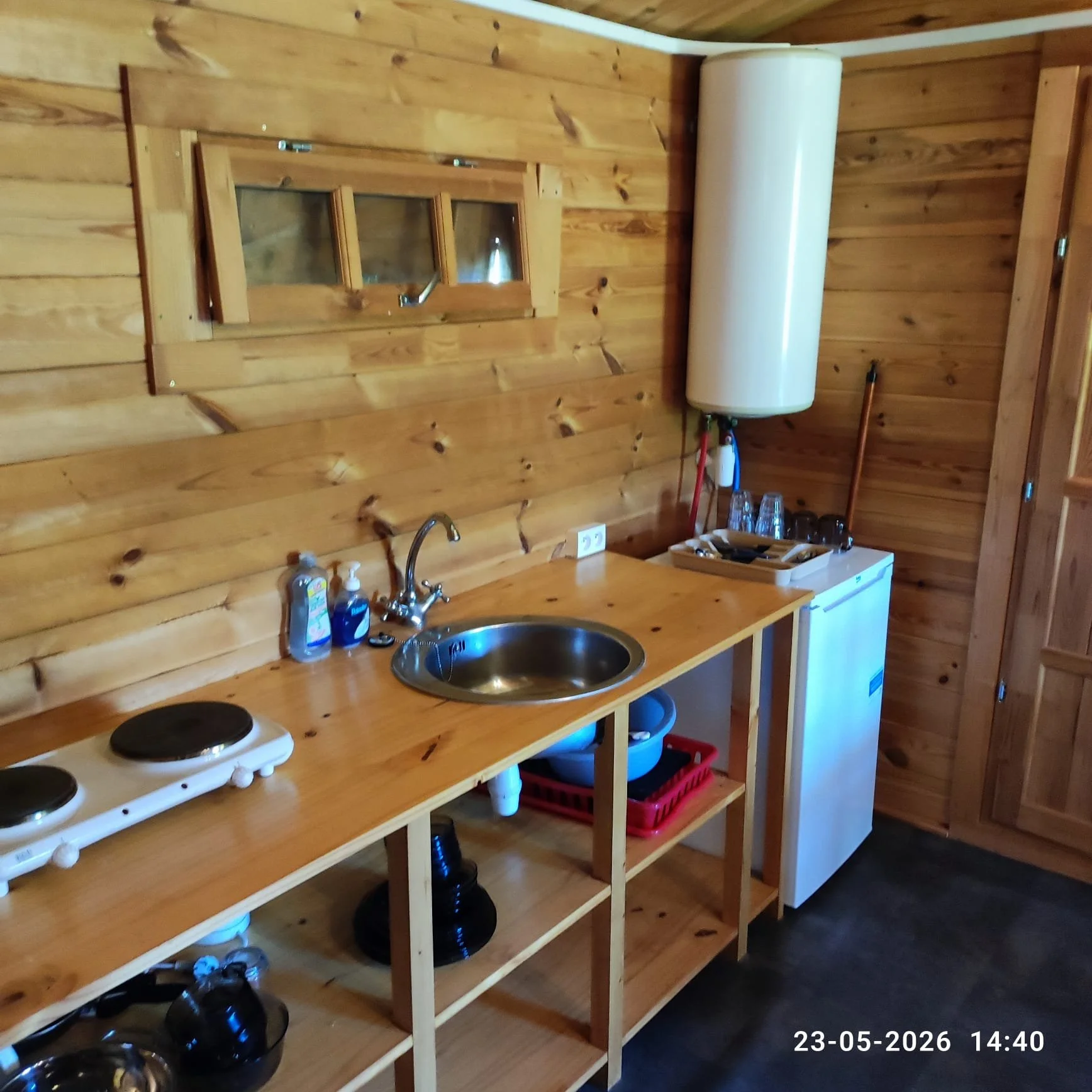 Een houten keukenkast met een wasbak, een inductiekookplaat, een kleine koelkast en keukengerei. Een waterboiler is aan de muur bevestigd. De klok onderaan de afbeelding toont de datum 23-05-2026 en tijd 14:40.