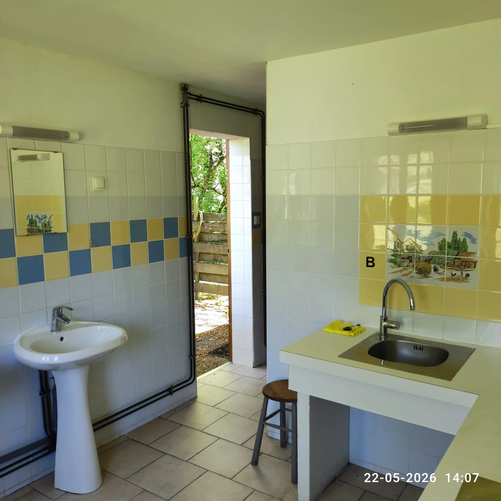 Binnenkant van een keuken met een wit tochttafel, een gootsteen, een spiegel en een klein decoratief tegelmuur. Een deur leidt naar buiten, waar bomen zichtbaar zijn. Datering: 22-05-2026, tijd: 14:07.