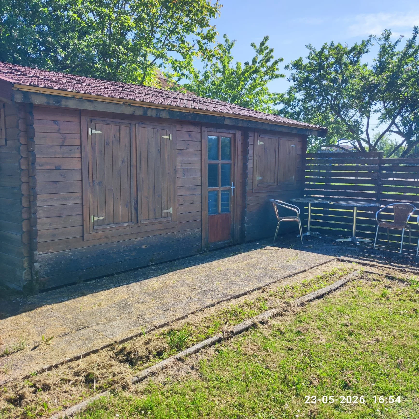 Een houten tuinhuisje met een dak bedekt met dakpannen, omringd door grote groene bomen. Op de voorgrond is een tuinpad te zien met enkele plantjes. In de rechteronderhoek staat de datum en tijd: 23-05-2026 16:54.