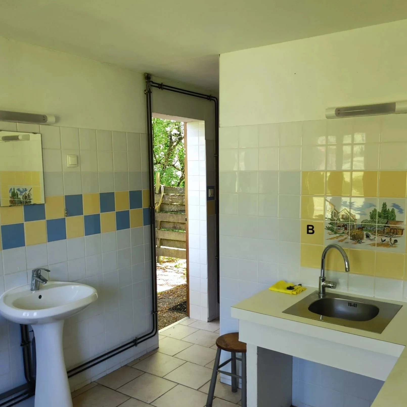 Een keuken met een wasbak, een muur met gekleurde tegels, een open deur naar buiten, een spiegel, een keukentafel met een kruk, en een gele doek met een kraan.