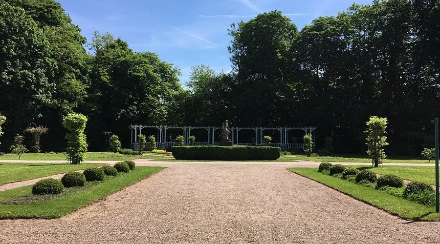 Een formele tuin met een zandpad dat naar een standbeeld in het midden leidt, omgeven door strak gesnoeide heggen en bomen onder een blauwe hemel.