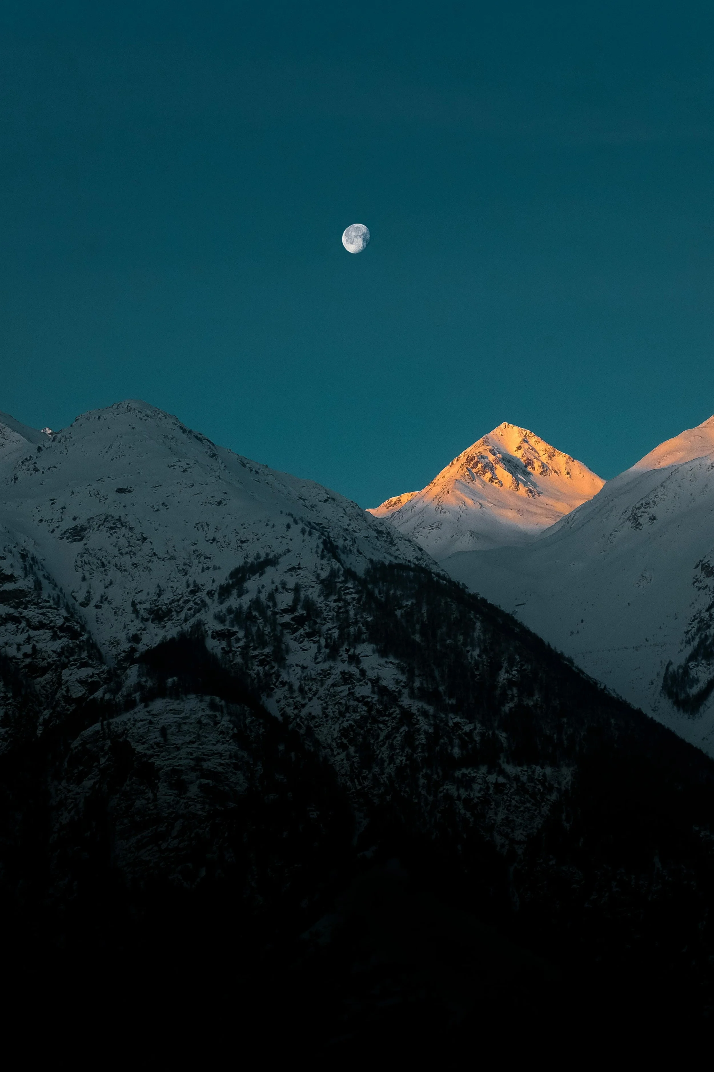 Snow-covered mountain peaks with a glowing orange hue from the setting or rising sun, under a clear sky with the moon visible.