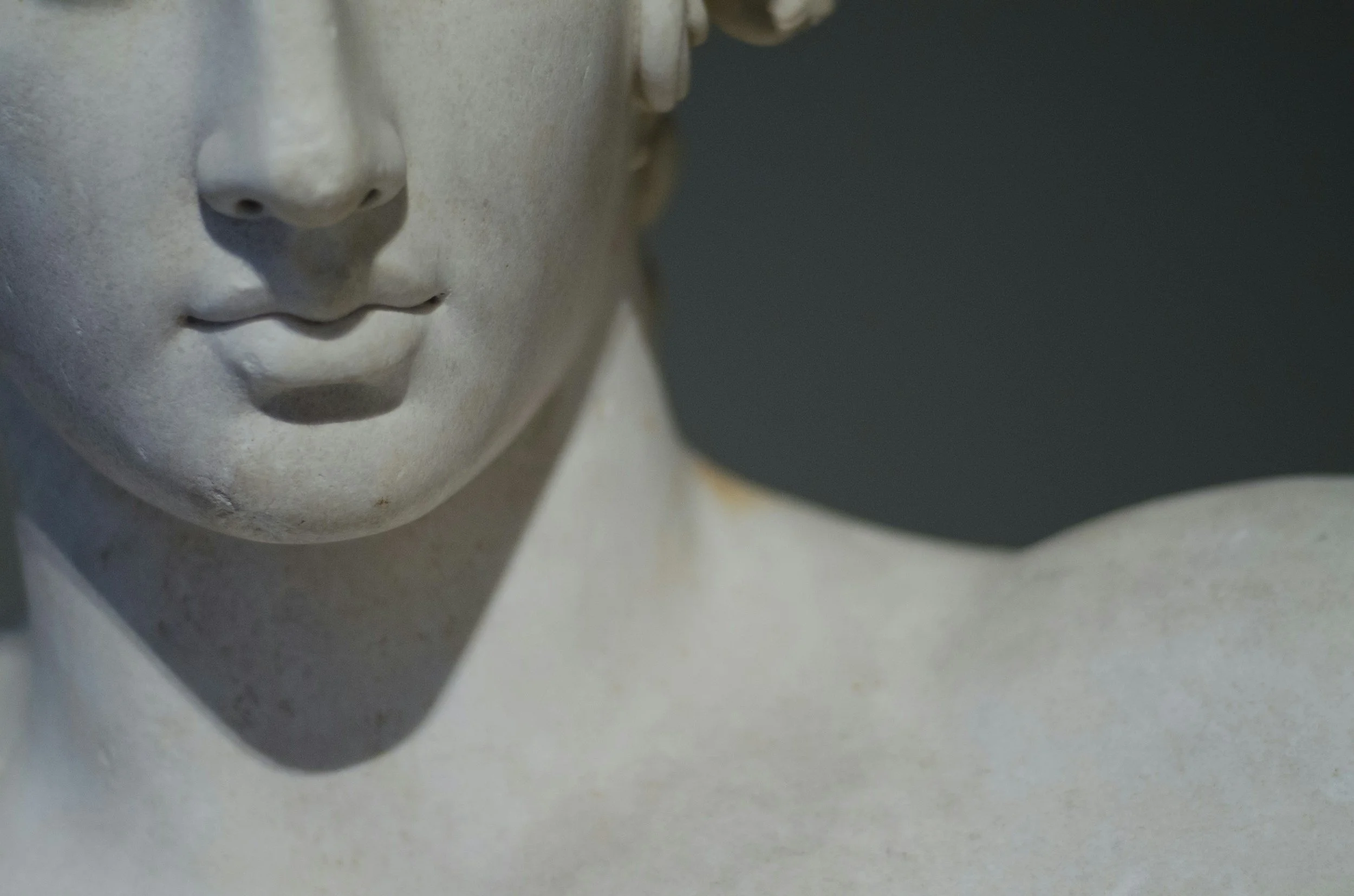 Close-up of a classical-style sculpture's face, highlighting the nose and lips, with a dark background.