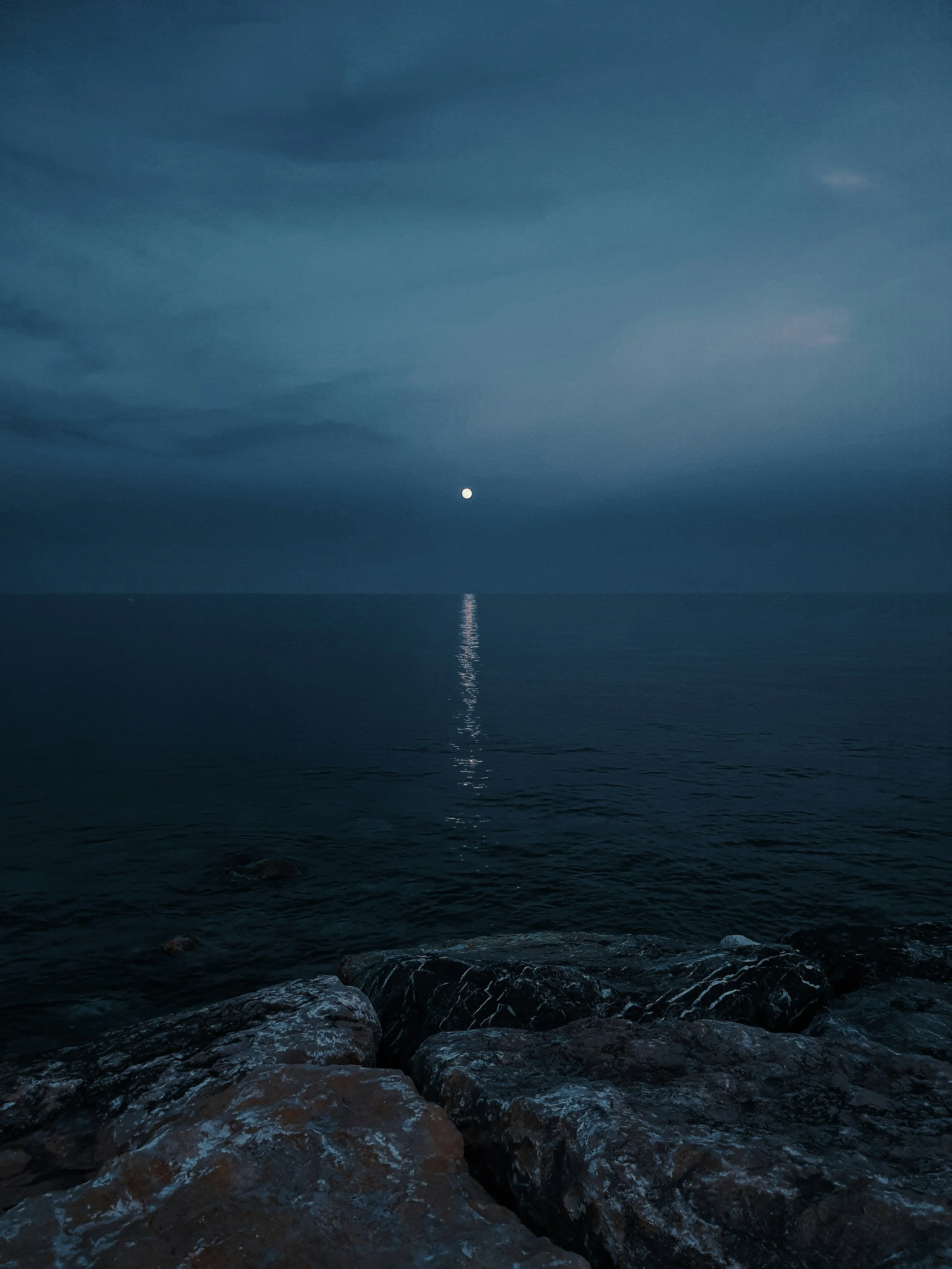Nighttime scene of the ocean with rocks in the foreground, a full moon reflecting on the water, and dark clouds in the sky.