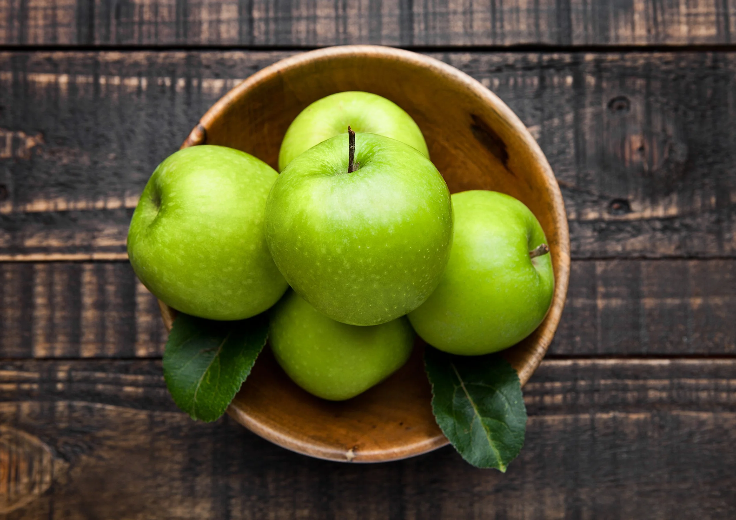 A small wooden bowl filled with green apples on a dark wooden surface.