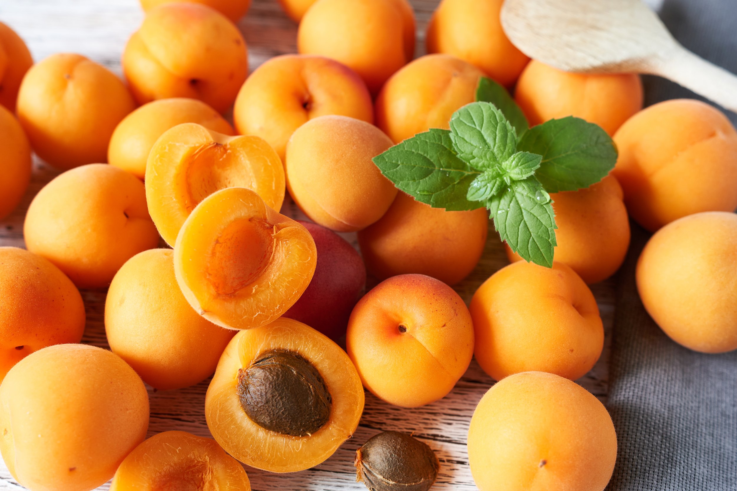 A collection of ripe apricots, some whole and some sliced open, with a sprig of fresh green mint on top. The apricots are on a wooden surface.