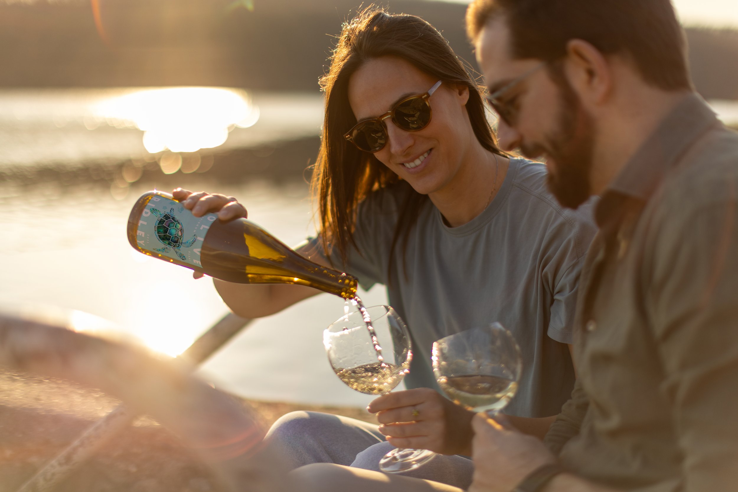 A smiling woman with long brown hair, wearing sunglasses, pours sparkling wine into a glass at sunset on the beach, accompanied by a bearded man with glasses.
