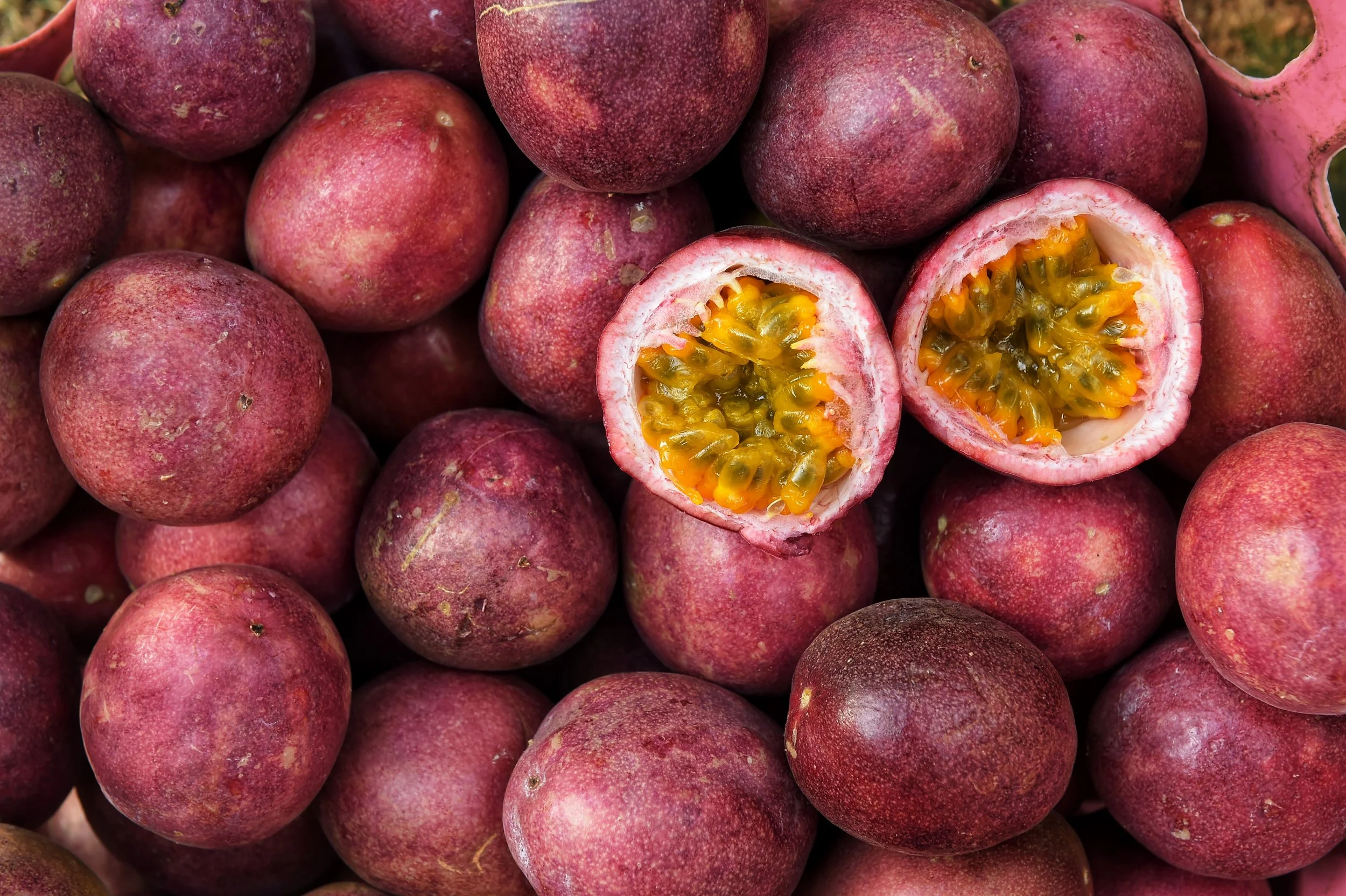 A pile of fresh passion fruits, with one cut open to show the yellow pulp and black seeds inside.