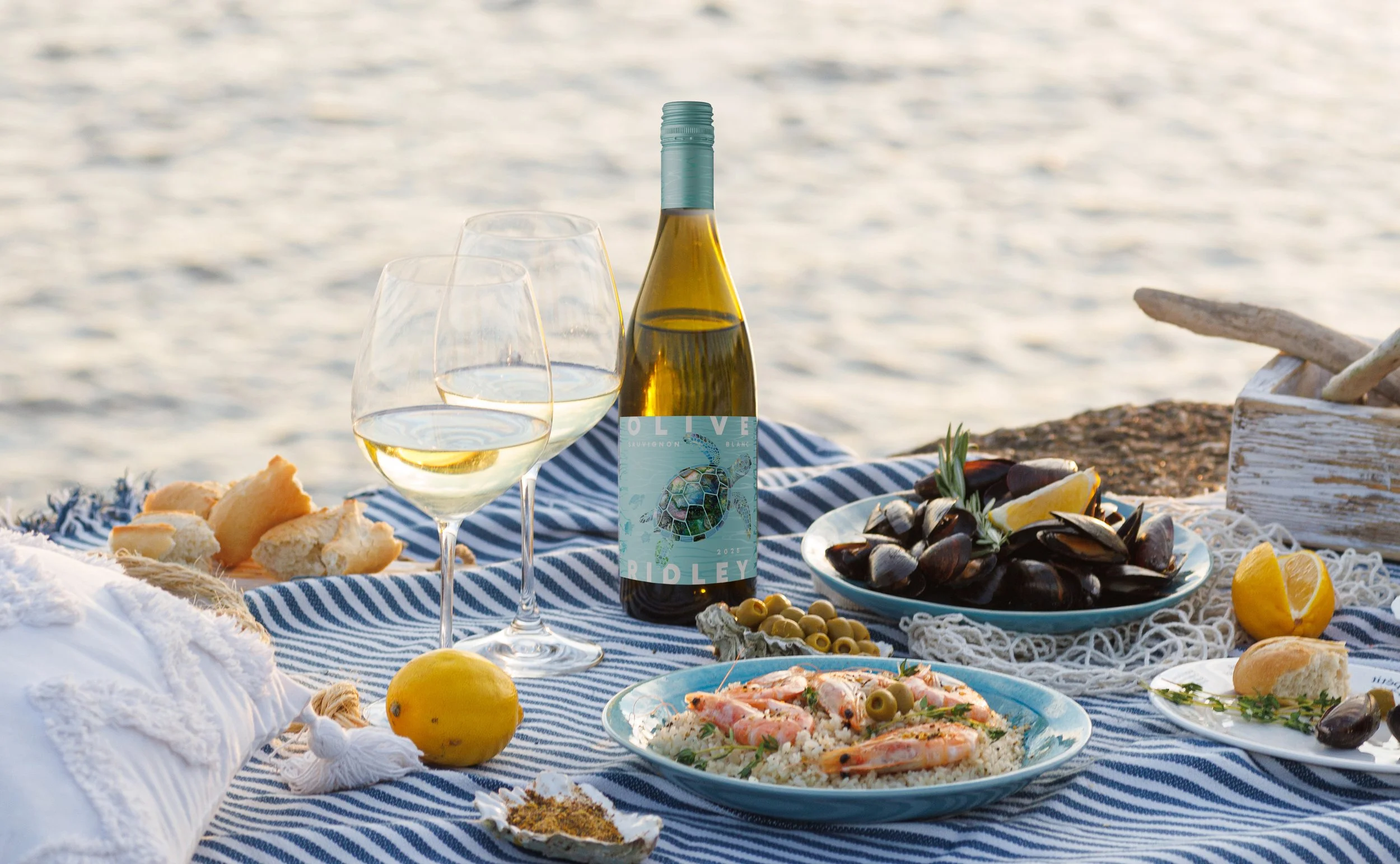 A seaside picnic setup with a bottle of white wine, two wine glasses, plates of seafood, bread, lemon, and other appetizers on a blue and white striped cloth near the water.