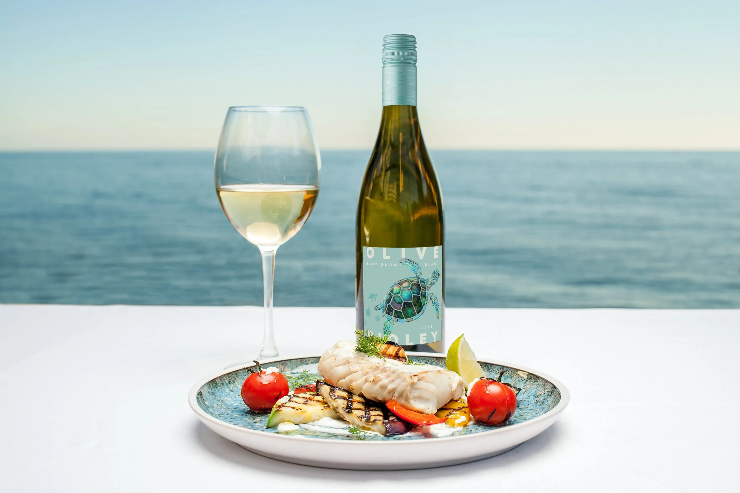 A plate of grilled seafood with cherry tomatoes, grilled vegetables, and a wedge of lime. Behind the plate, there is a bottle of white wine with a sea turtle illustration and a glass of white wine. The background shows the ocean under a clear sky.