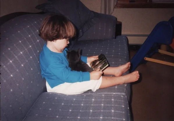 Leah as a toddler reading a book with a cat in her lap