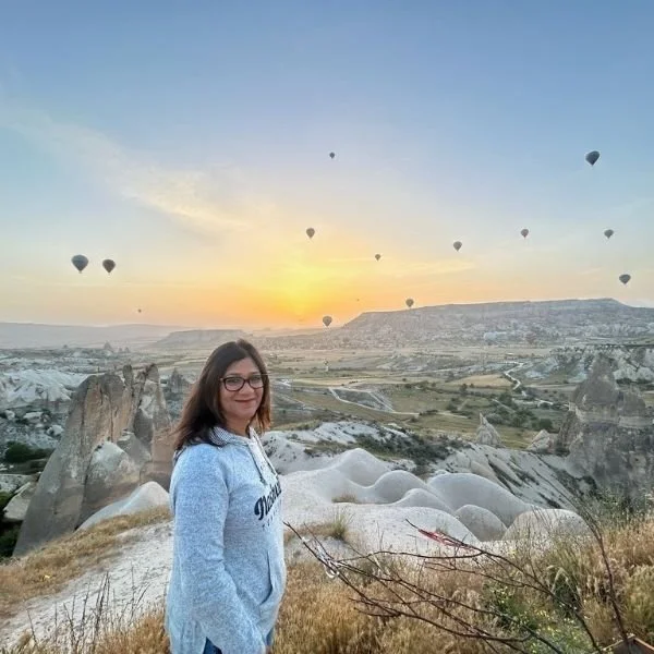 Monica Maheshwari smiling at sunset in a landscape with rock formations and hot air balloons in the sky.