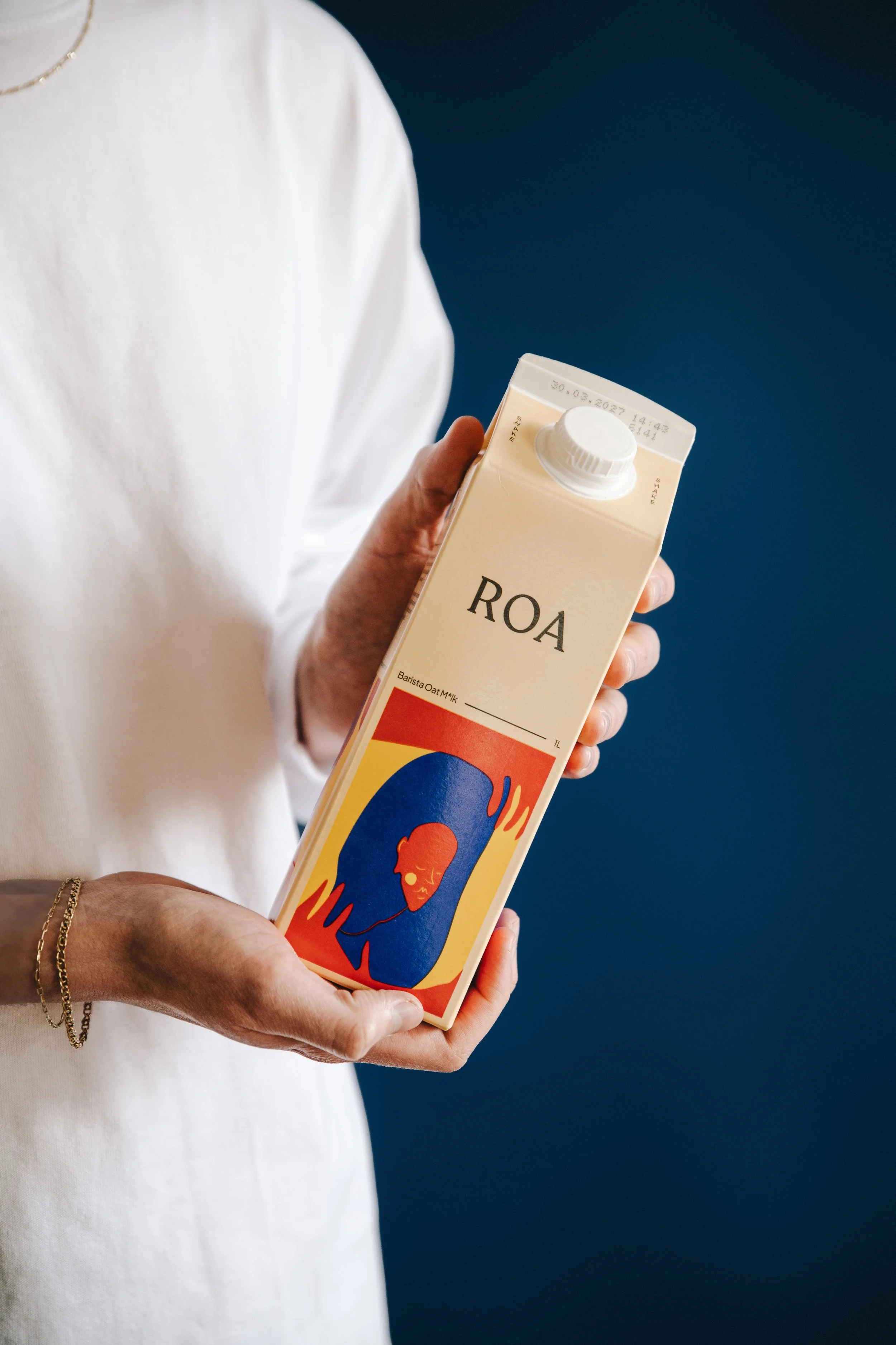 Person holding a carton of ROA oat milk with a colorful artistic label featuring a stylized face with flowing hair and flames, wearing a white long-sleeved shirt, against a dark blue background.
