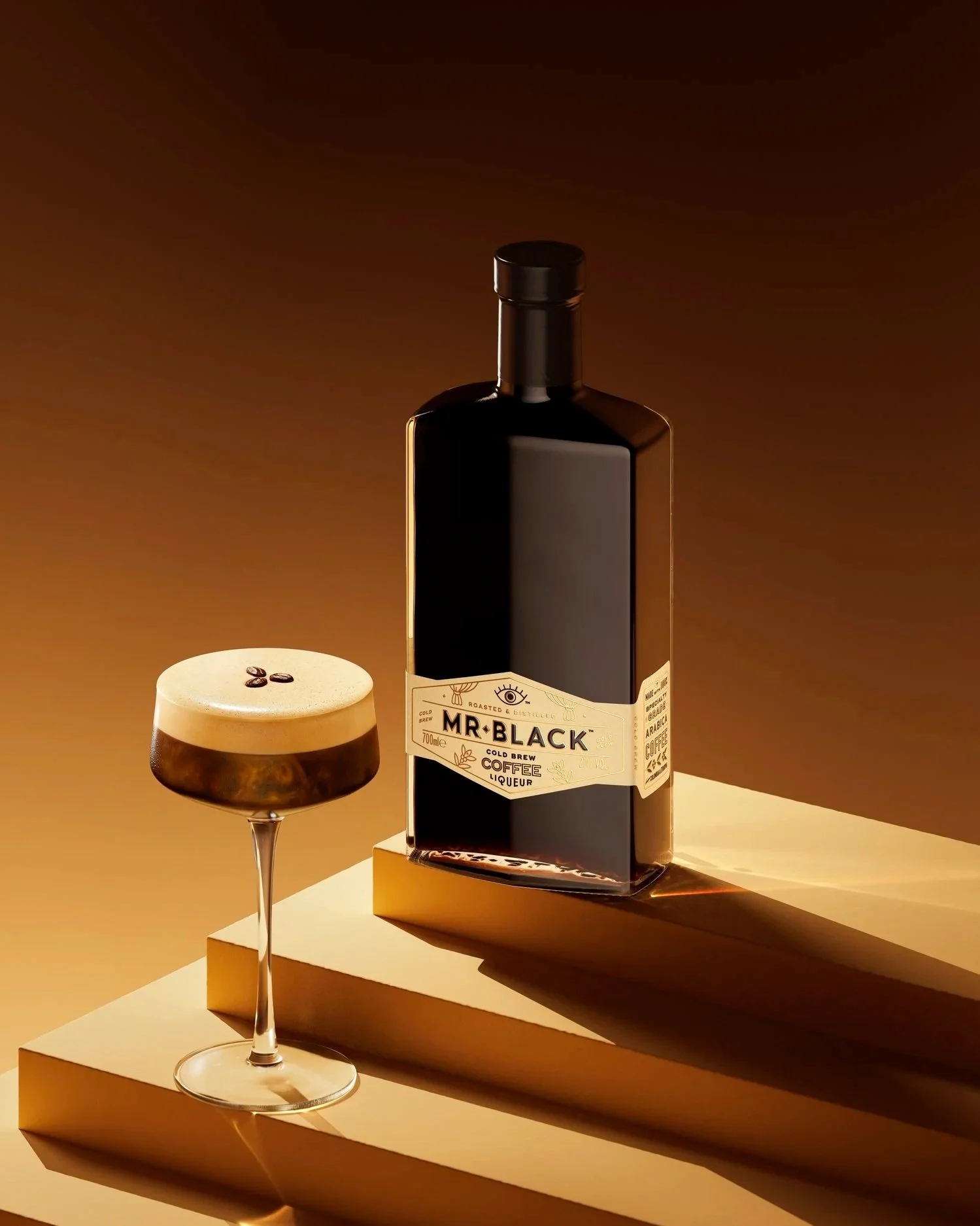 A dark bottle of Mr. Black coffee liqueur and a glass of iced coffee with ice cubes and coffee beans on top, placed on a stepped white surface with warm lighting creating shadows.