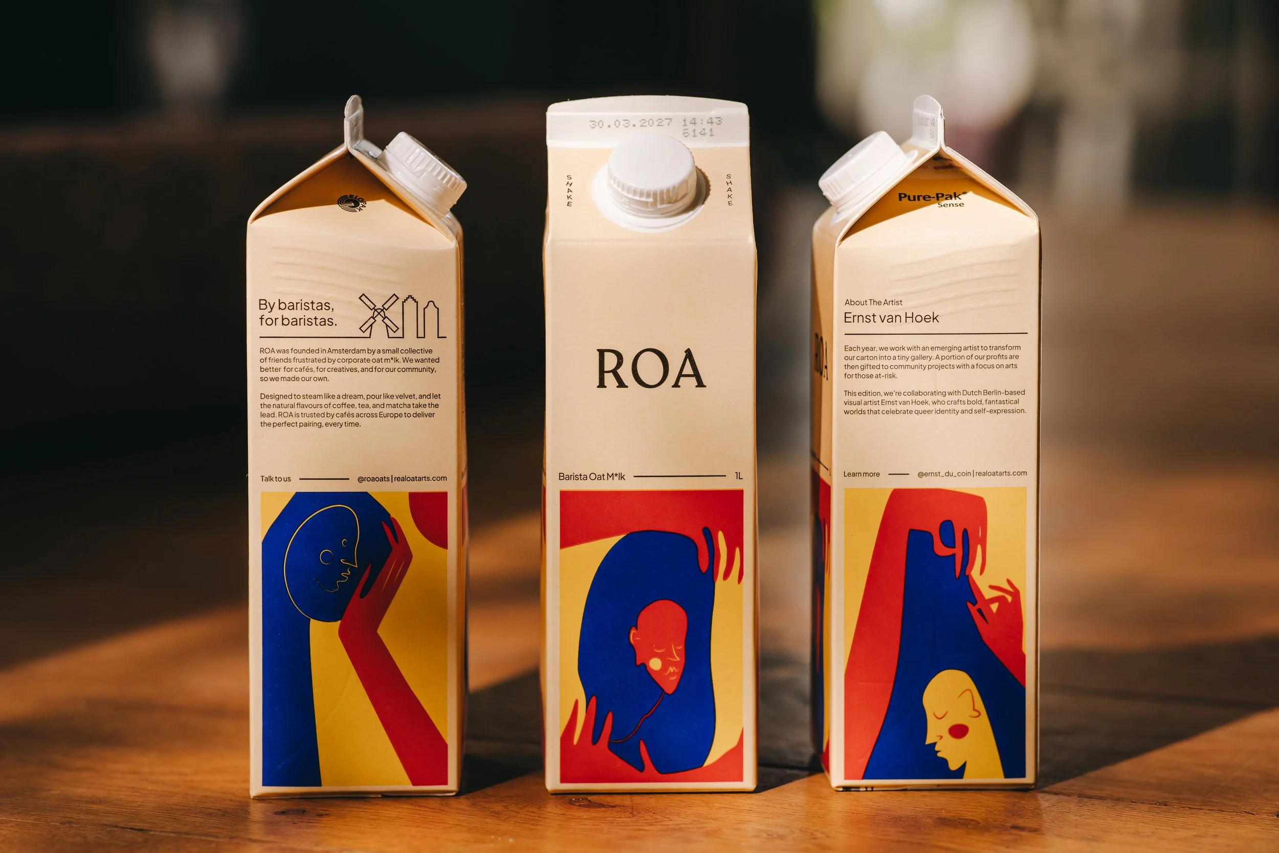 Three cartons of Oatly oat milk on a wooden surface with a dark background, each featuring a colorful abstract human face illustration.