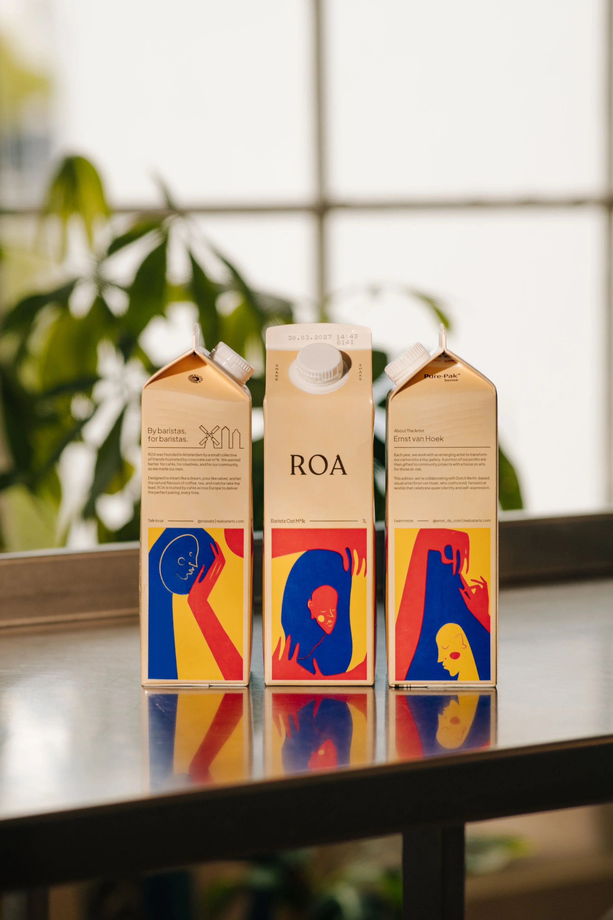 Three cartons of ROA oat milk on a table with a blurred green plant in the background and a window letting in light.