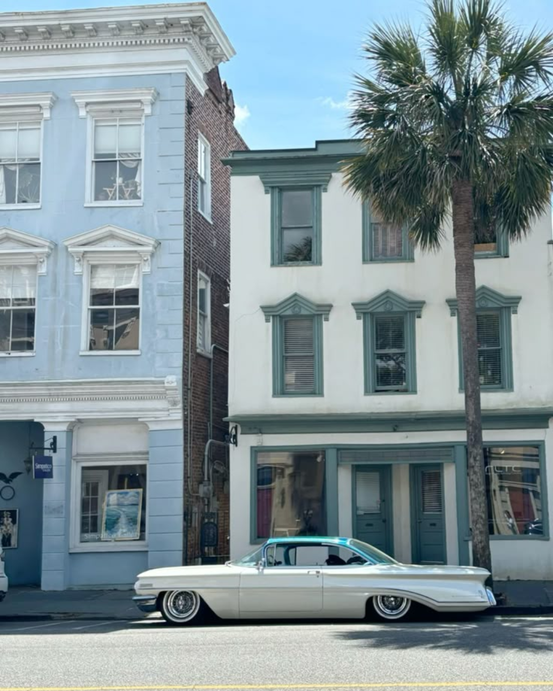 A vintage white car parked on a street in front of two-story buildings with large windows and decorative trim, one building is painted light blue and the other is white with teal accents, a palm tree is visible to the right.