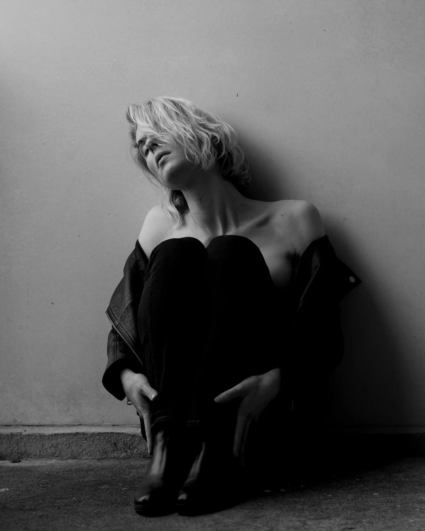 Black and white photo of Elsa Léger with short, wavy hair, topless, wearing a jacket off the shoulders, sitting against a plain wall with her head tilted back and eyes closed.