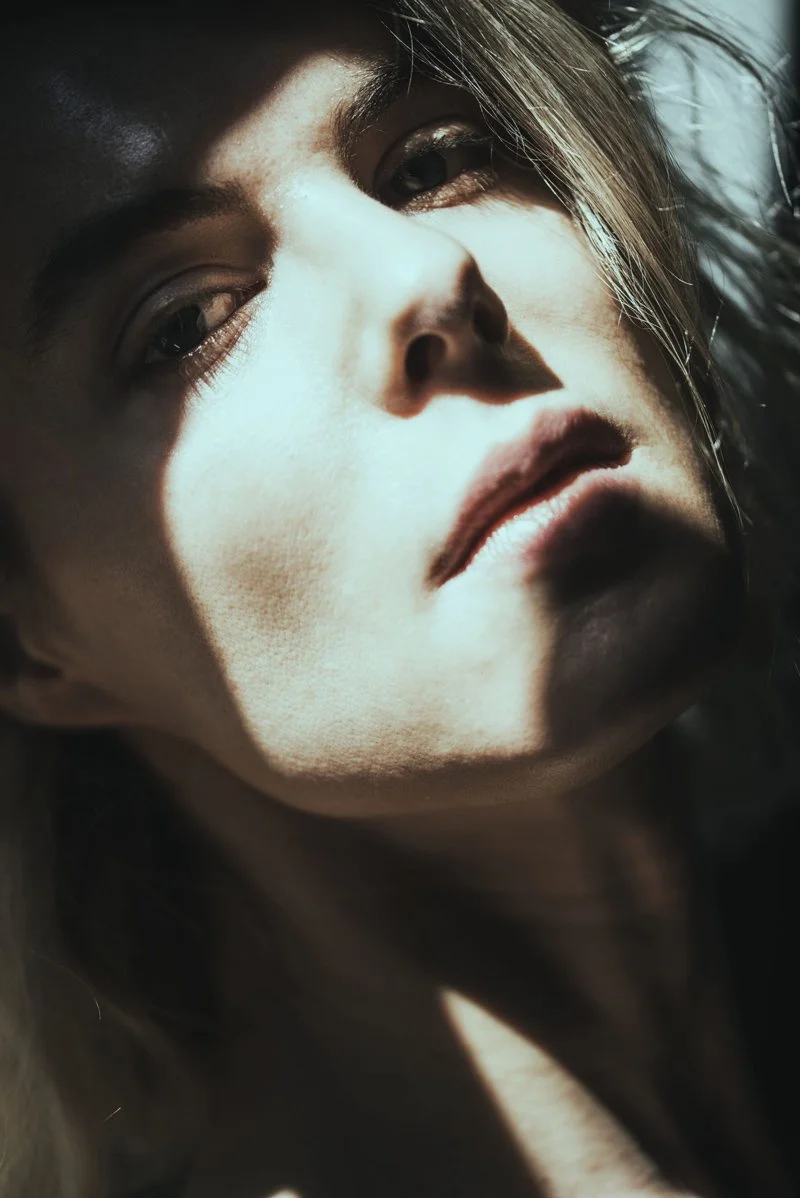 Close-up of Elsa Léger's face with dramatic shadows and lighting, partially illuminated from above, creating high contrast and emphasizing facial features.