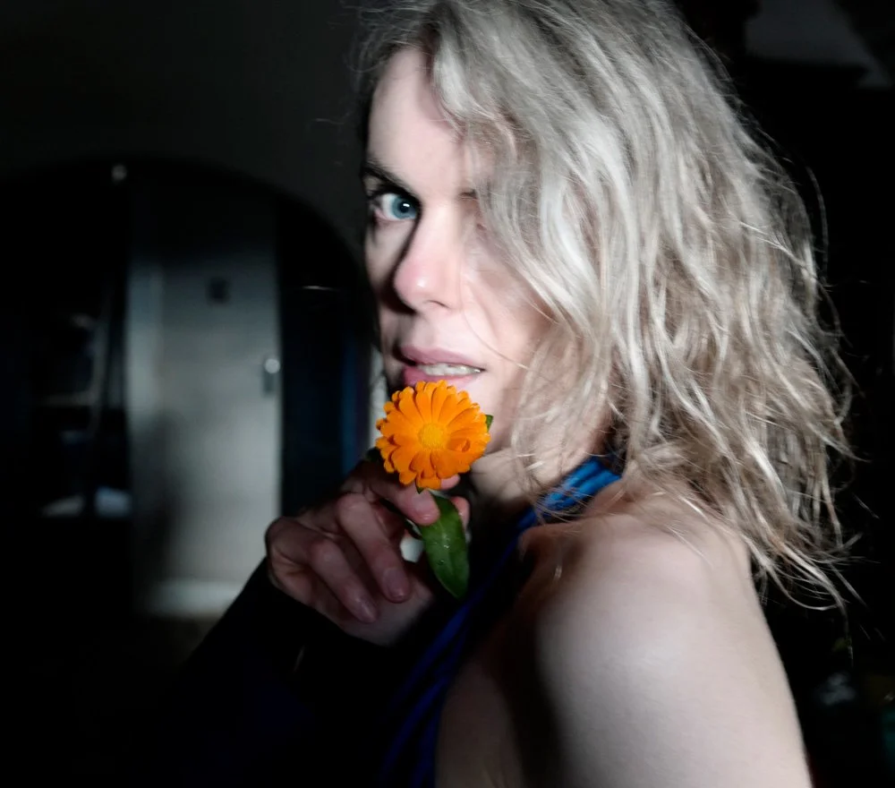 Elsa Léger with curly blonde hair and blue eyes holding an orange flower near her mouth, looking at the camera.