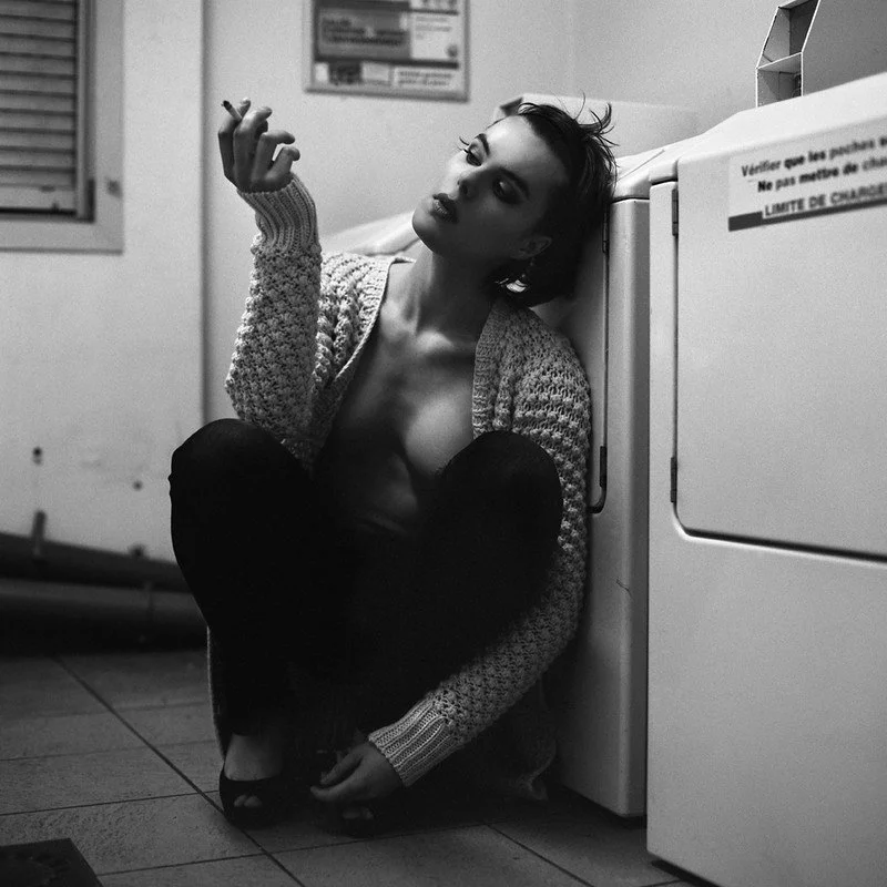 Elsa Léger sitting on the floor in a laundromat, leaning against a washing machine, wearing a knitted cardigan and dark pants, with short hair, looking thoughtfully at her hand.