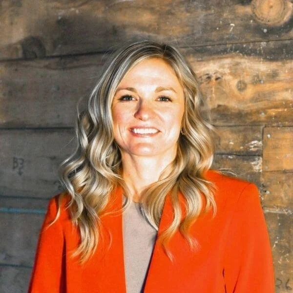 A woman with long blonde wavy hair smiling, wearing a red blazer over a beige top, standing in front of a wooden wall background.