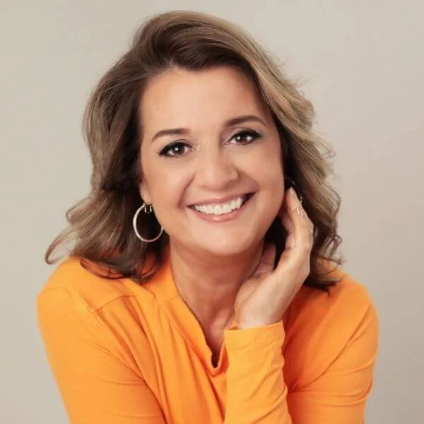A woman with wavy light brown hair, wearing a yellow top and hoop earrings, smiling and touching her face with one hand.