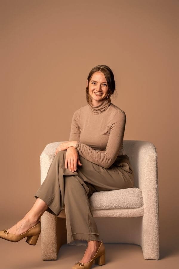 Headshot Tess Hobson, CEO of The Story Within  - smiling sitting on a light-colored sofa against a plain beige background, wearing a beige turtleneck, beige wide-leg pants, and beige high heels.