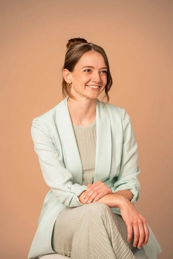 Headshot Tess Hobson, CEO of The Story Within - wearing earrings, a light green blazer, and a beige dress, smiling while seated against a beige background.