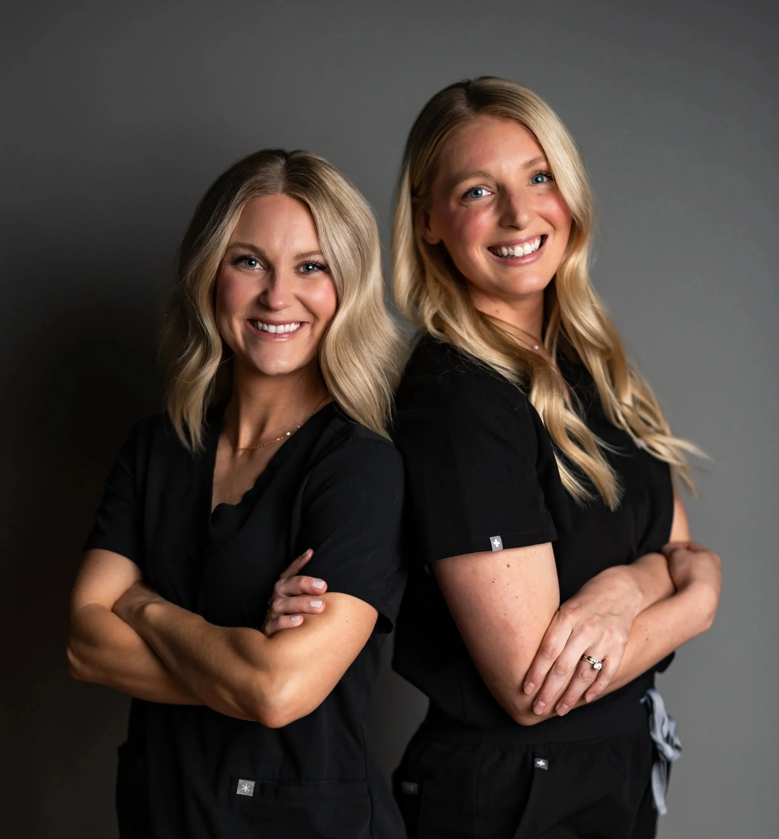 Two women with blonde hair smiling and hugging each other against a gray background.