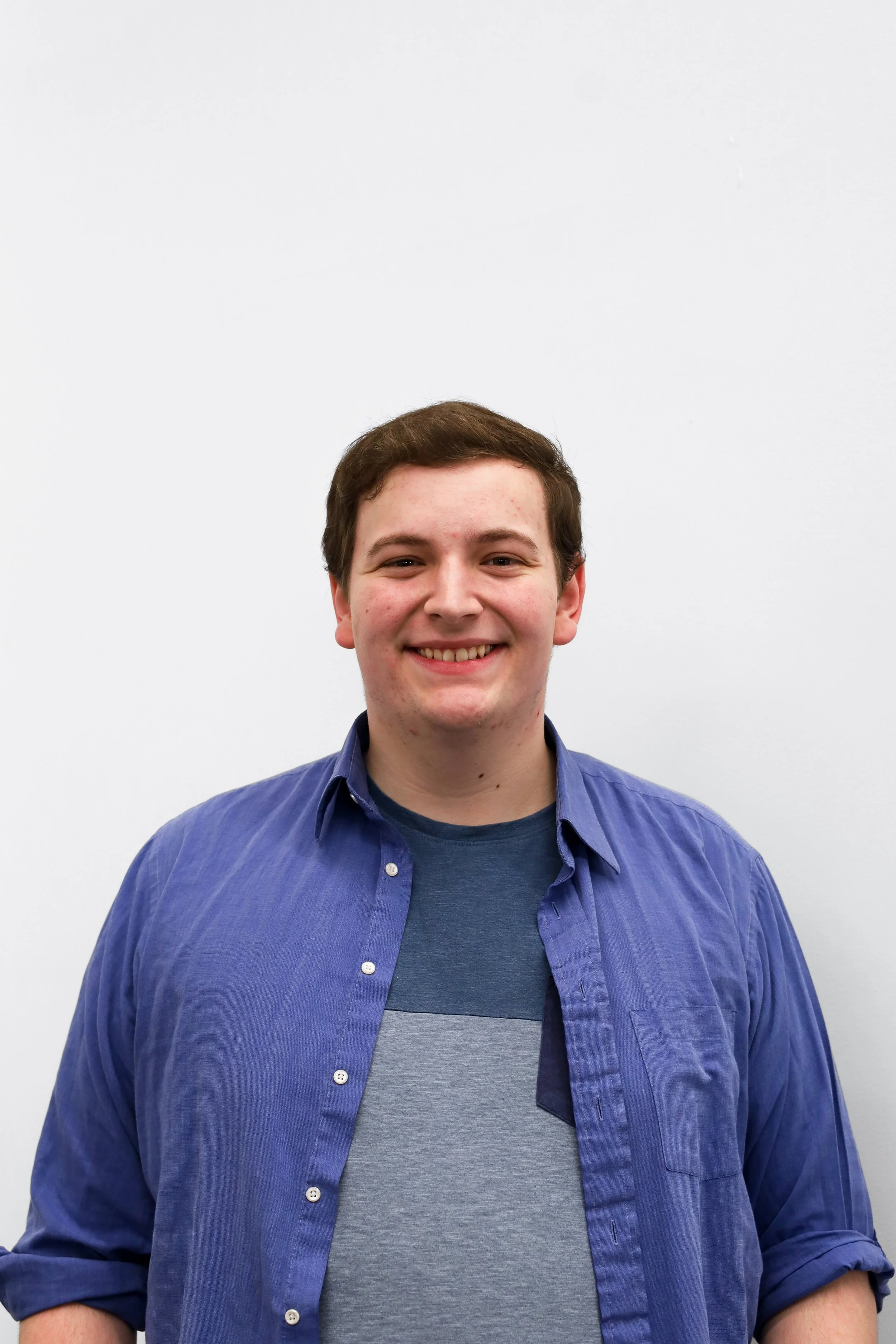 Headshot of Nathan Lanum wearing a blue dress shirt and a blue undershirt against a white background, smiling