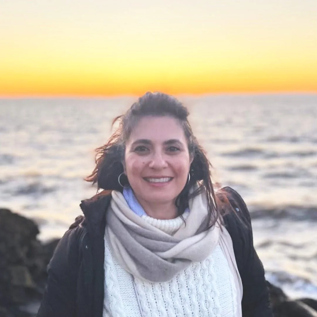 Smiling woman with hoop earrings, wearing a white sweater and scarf, standing near rocks by the ocean at sunset.