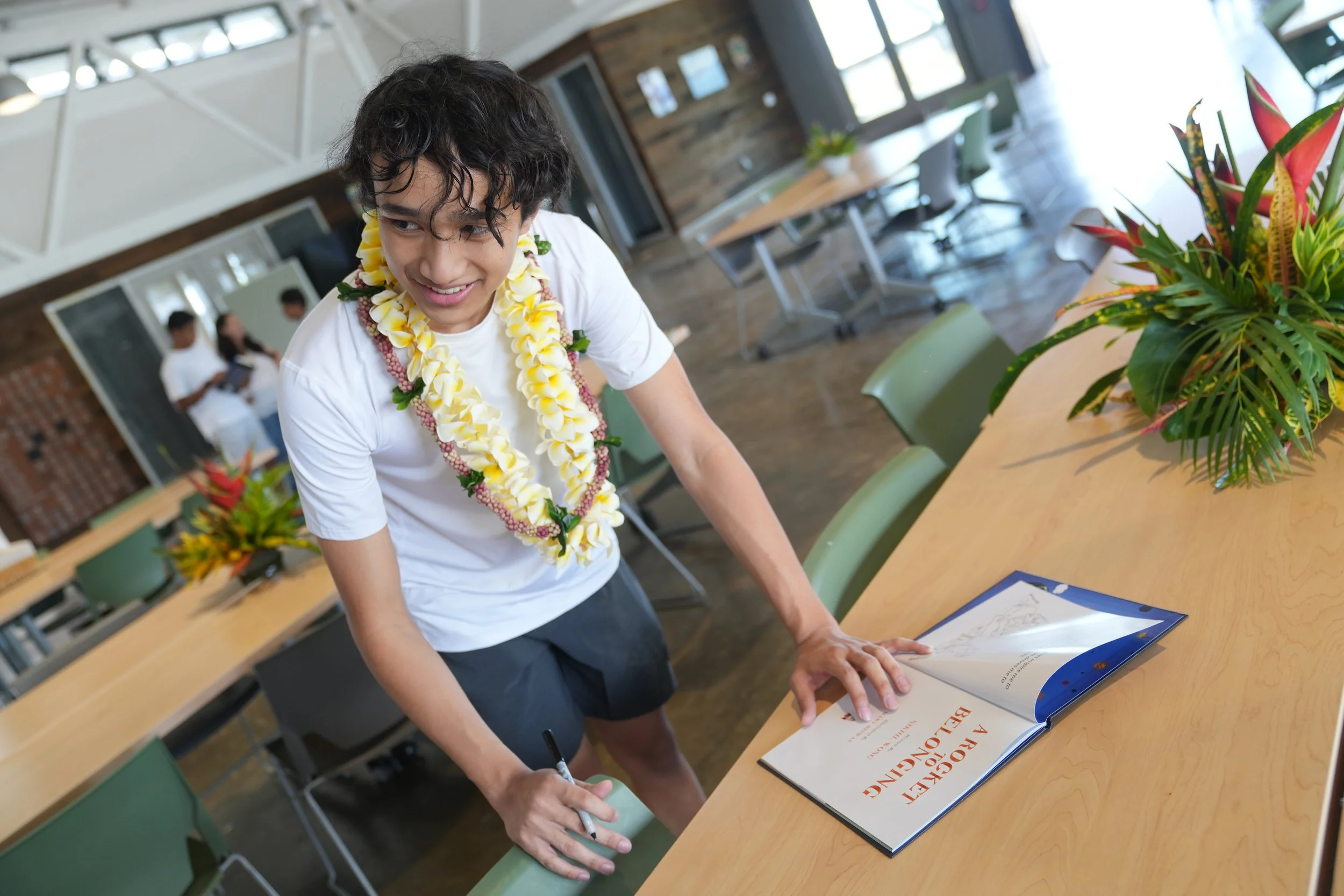 A young man wearing a white shirt and floral lei is signing a document on a table in a modern office space with large windows and wooden walls.