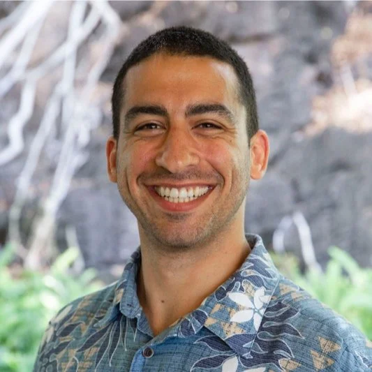 A smiling man with short dark hair wearing a patterned blue button-up shirt, outdoors with blurred greenery and rocks in the background.