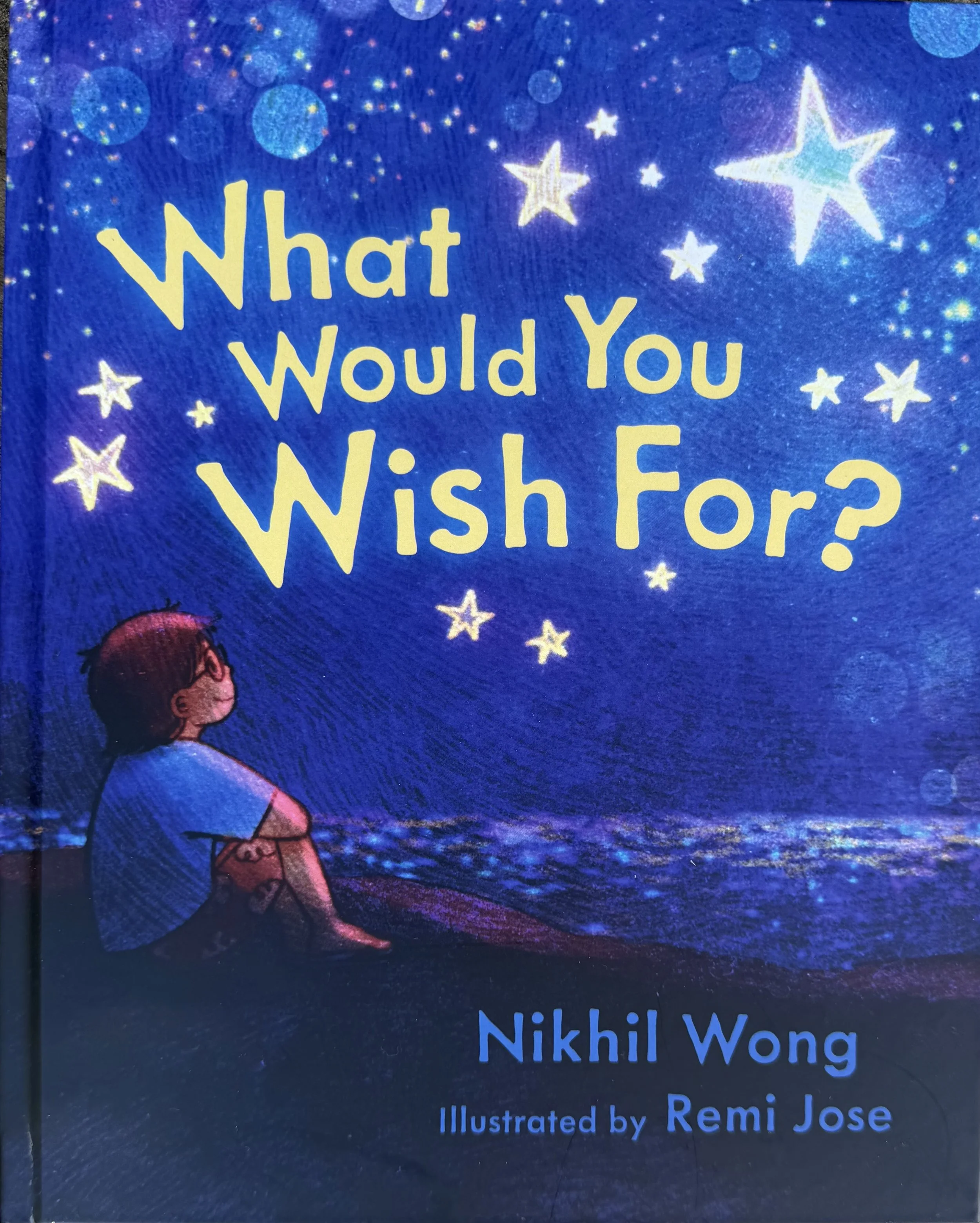 Cover of a children's book titled "What Would You Wish For?" with a starry night sky sky background and an illustration of a girl sitting on a dock by water. The text also includes the author Nikhil Wong and illustrator Remi Jose.