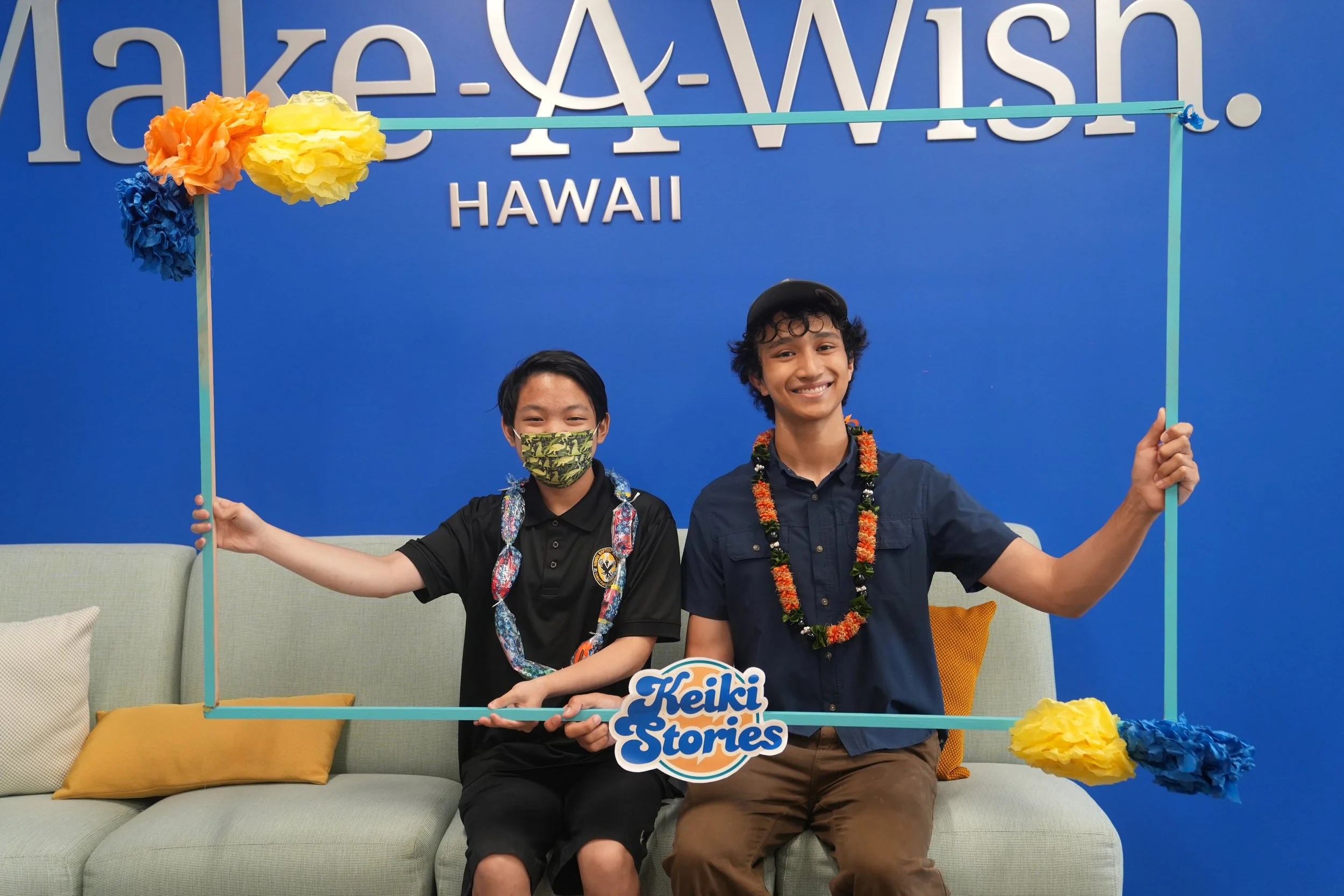 Two young men in Hawaii, one wearing a camo face mask, the other wearing a lei, holding a photo frame with the text 'Keiki & Stories' at an indoor event with a blue background and decorative flowers.