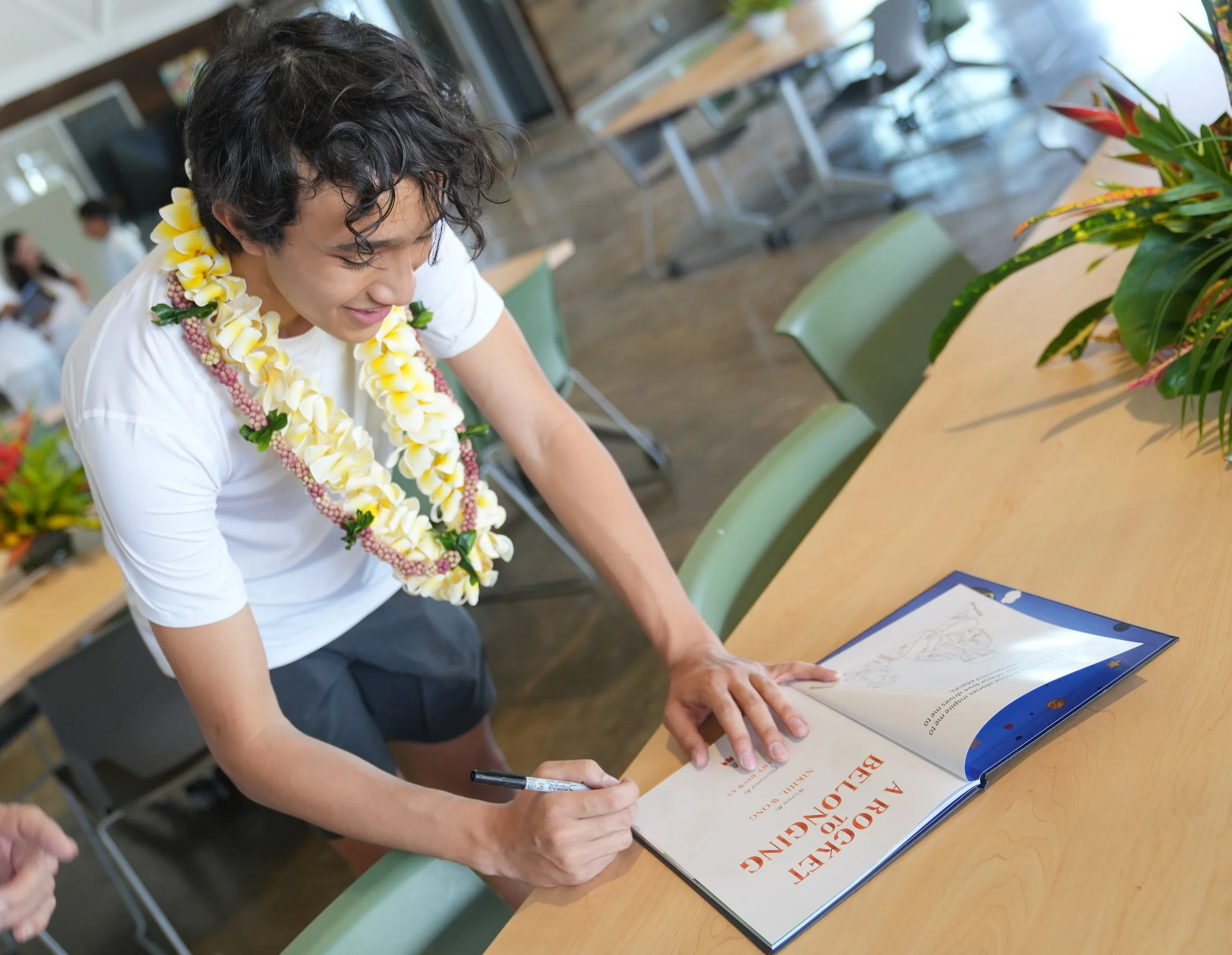 A teen boy with short curly hair, wearing a white shirt with a yellow floral lei, signs a book on a wooden table in a room with chairs and a plant nearby, in a casual or celebratory setting.