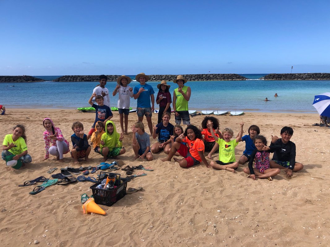 Group of children and adults on a sandy beach with ocean and clear blue sky in the background. Some people are in the water, and there are kayaks and beach gear. The group is smiling and posing for the photo.