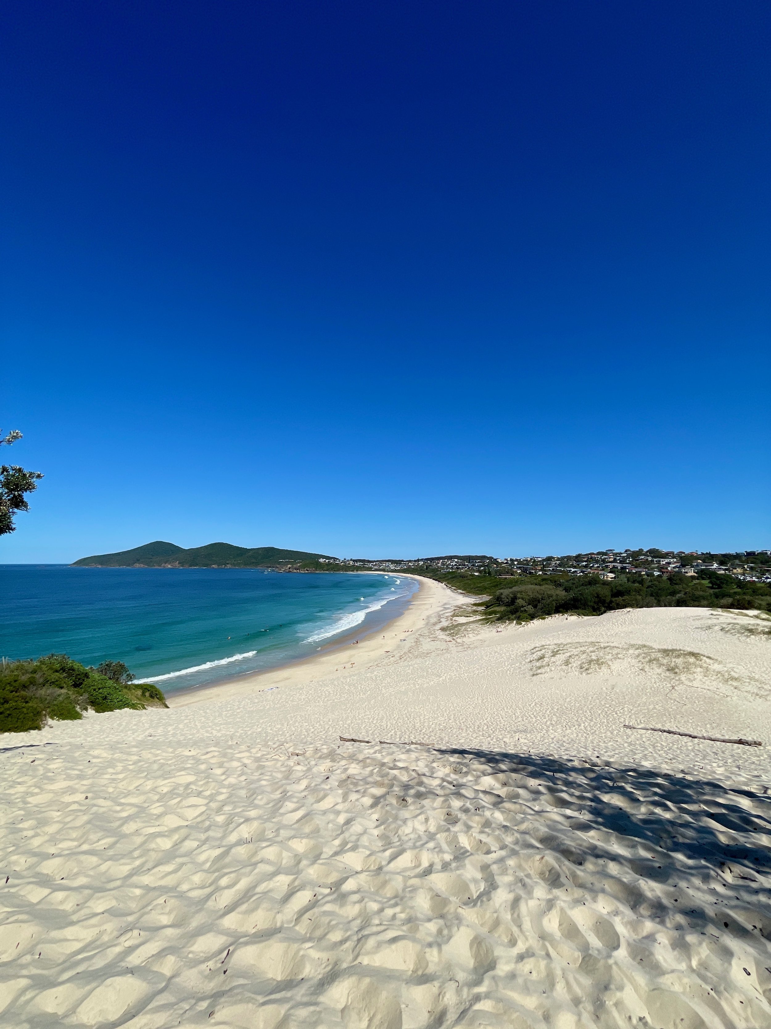 A scenic view of a sandy beach with turquoise water, a distant shoreline with houses and green hills, under a clear blue sky.
