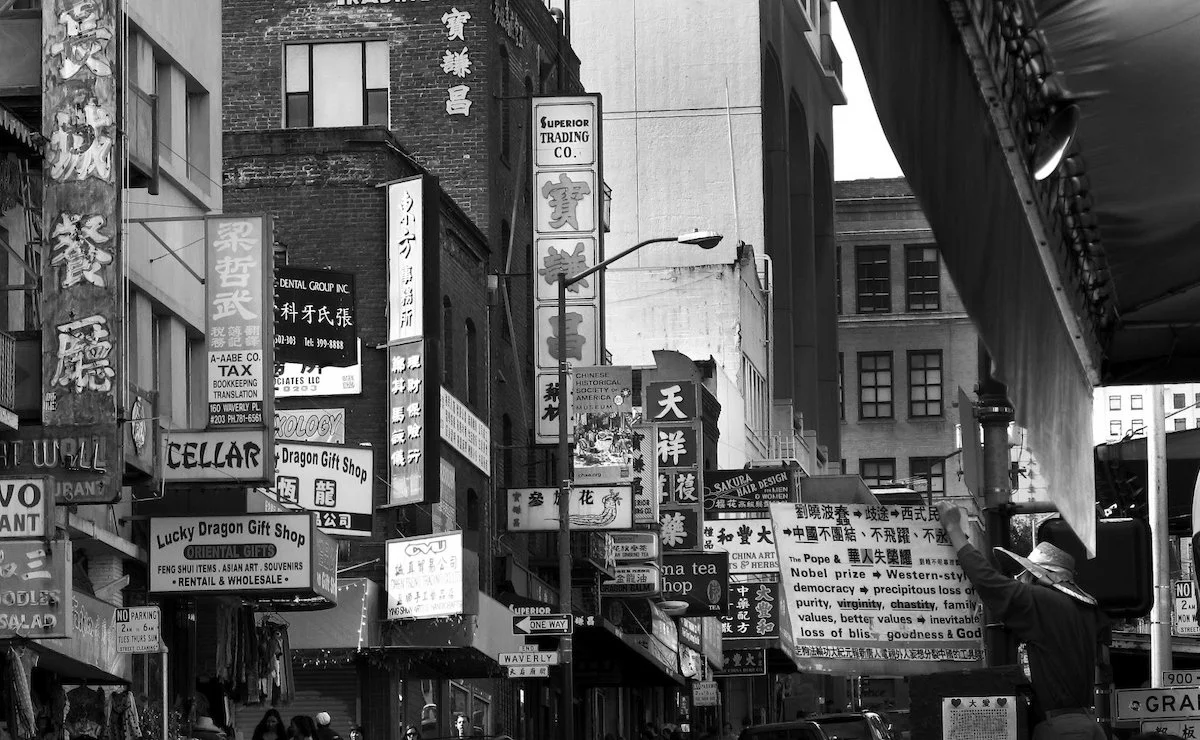 A black and white photo of a busy street with numerous Asian businesses and signage, including a gift shop, tea shop, and medical clinic, in a Chinatown area.