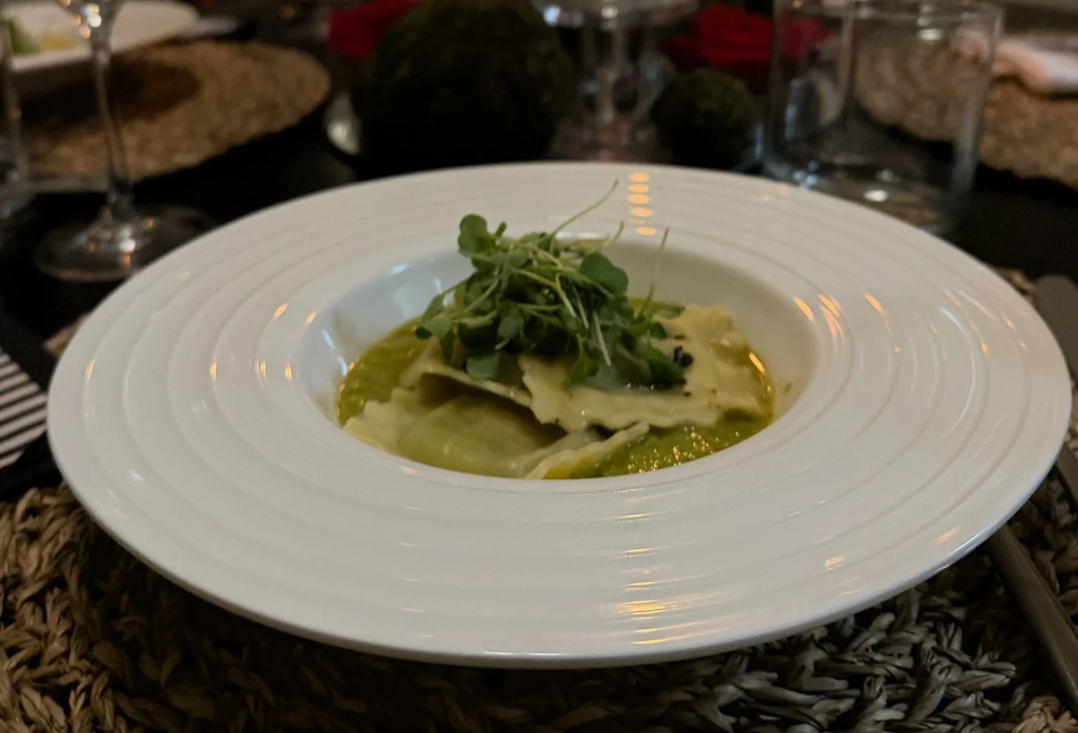 A plate of ravioli topped with microgreens, served with green sauce, on a white deep dish plate. In the background, wine glasses and a table setting are visible.