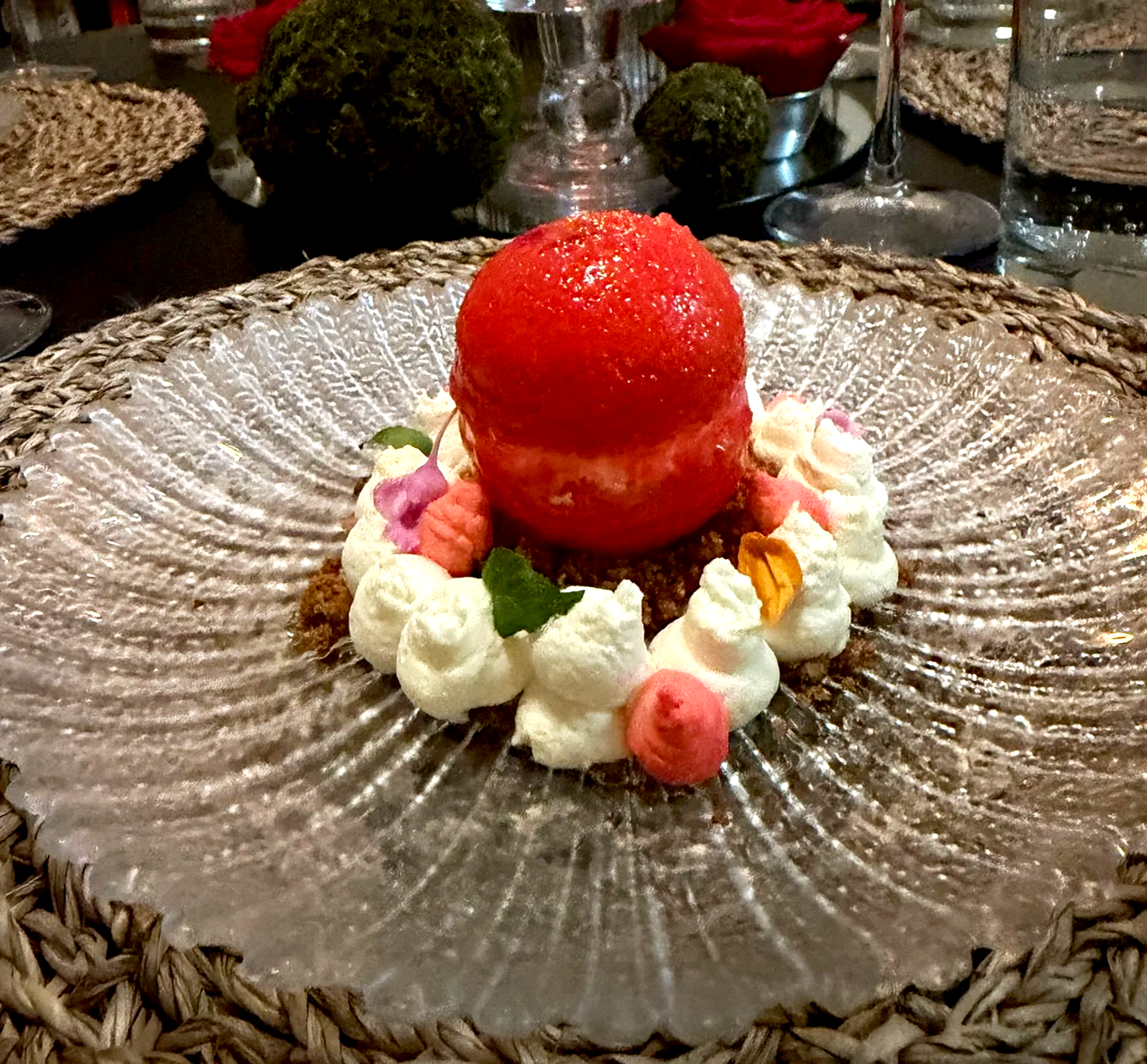 A dessert featuring a red sorbet or ice cream ball on a bed of whipped cream, garnished with edible flowers and mint leaves, served on a textured glass plate.