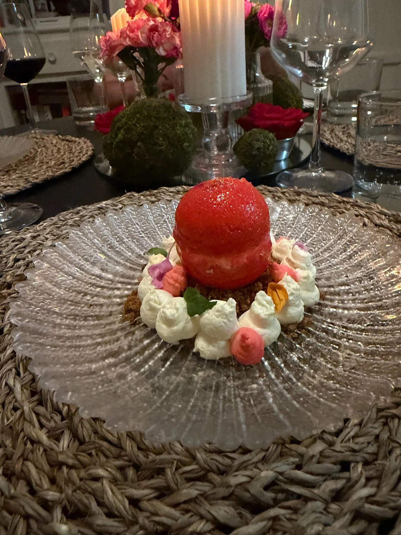 A dessert with a red spherical topping surrounded by whipped cream and colorful edible flowers, served on a textured clear glass plate with a woven placemat underneath. In the background, a candle, floral arrangements, and wine glasses are visible on a table.