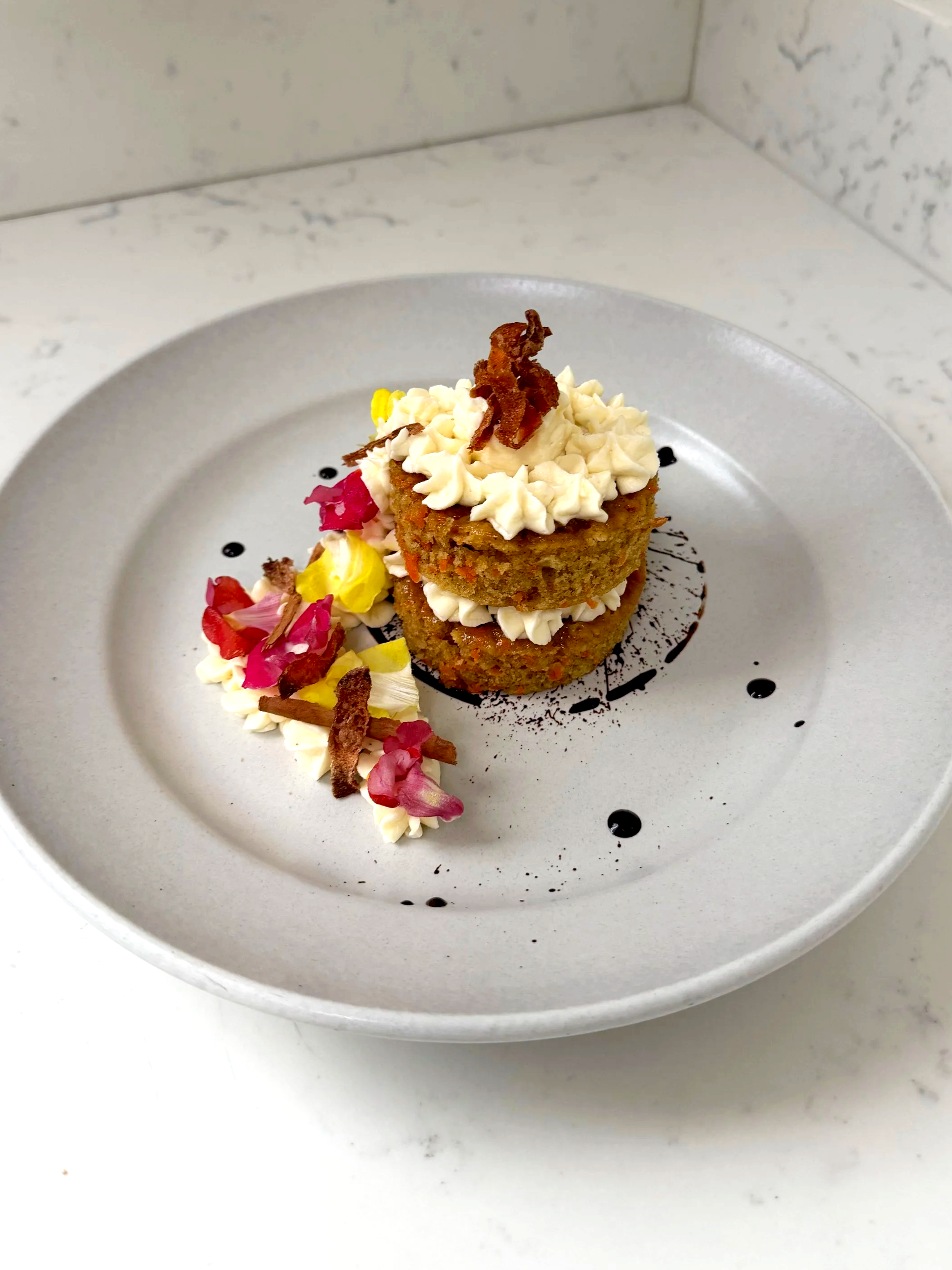 Two stacked breaded and fried rounds topped with cream, bacon, and herbs, served on a gray plate with decorative sauce and flower petals.