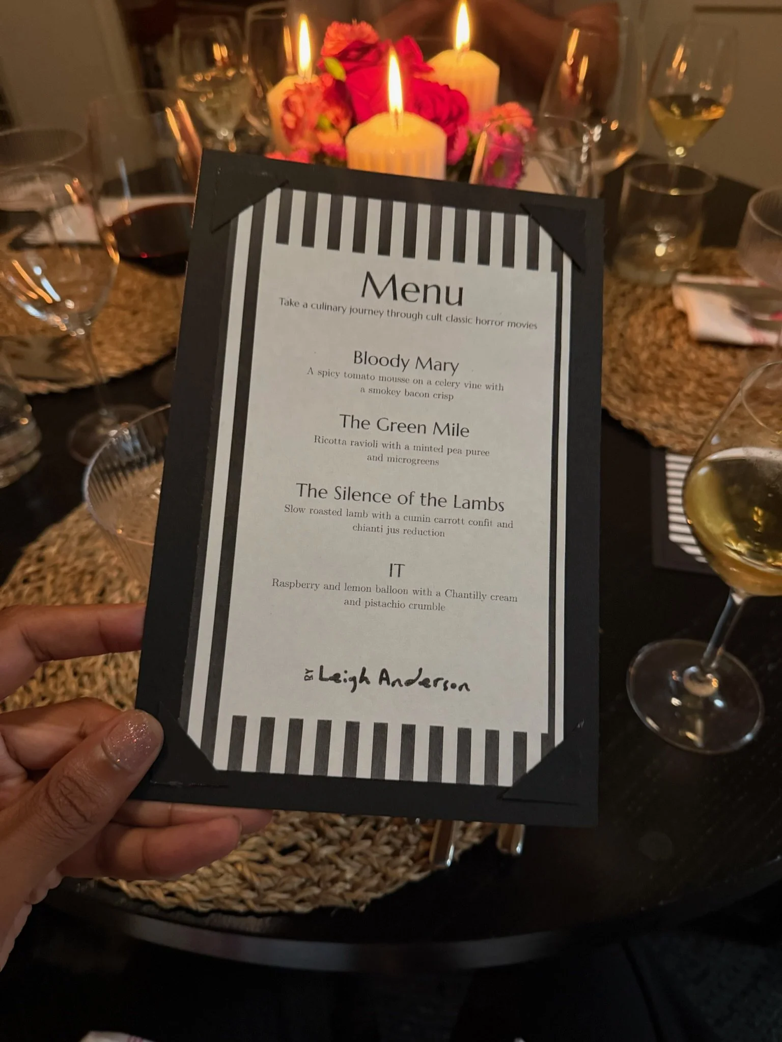 A menu card held in a person's hand in front of a table setting with wine glasses and a floral centerpiece with candles.