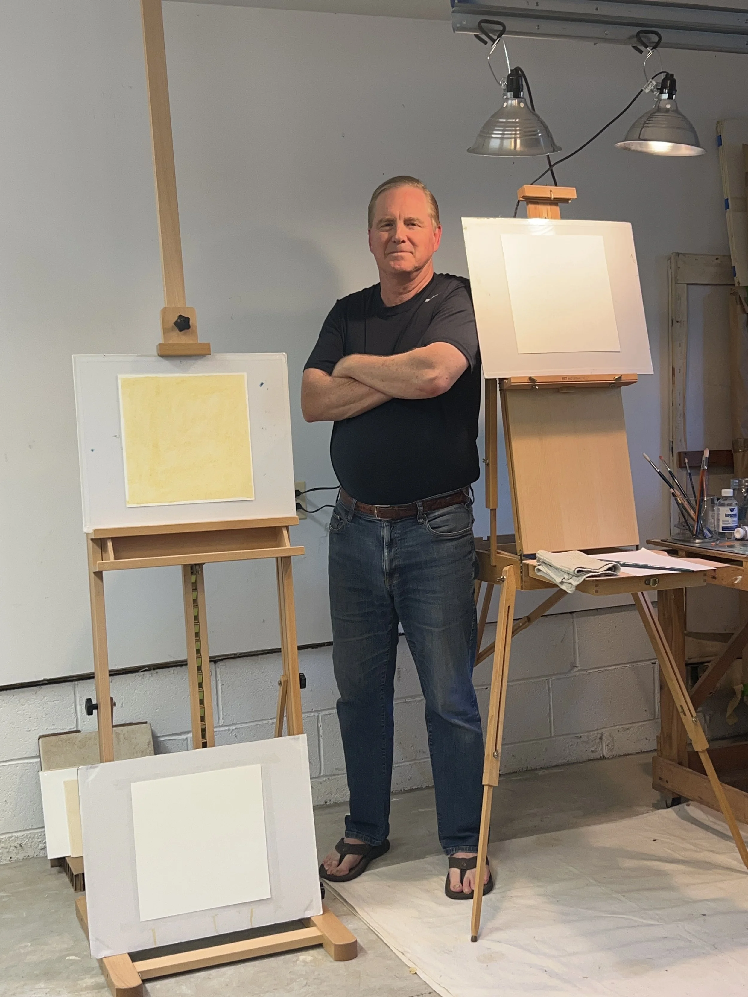 A man standing with his arms crossed in an art studio, surrounded by blank canvases and painting supplies, wearing a black shirt, jeans, and sandals.