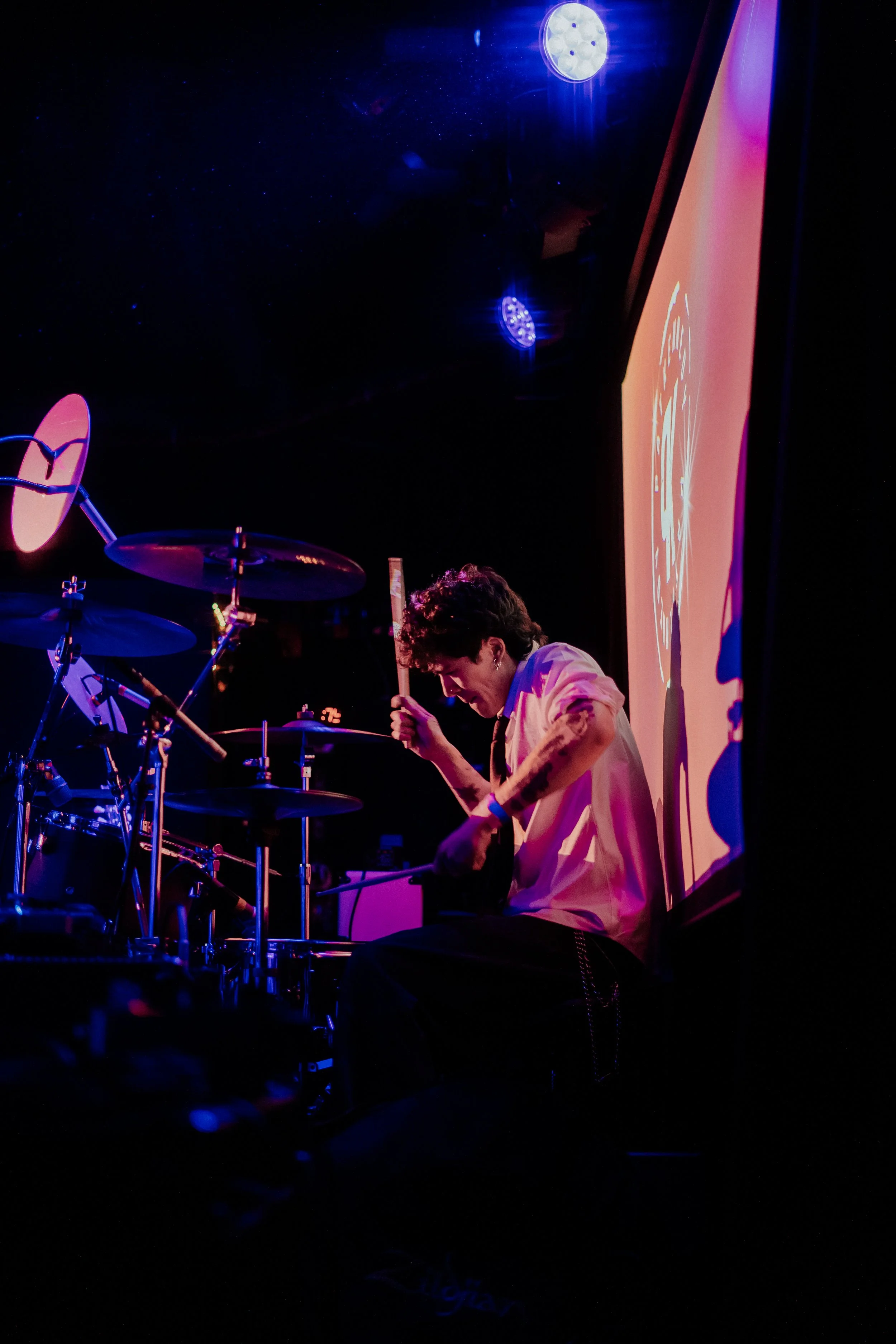 A person with dark curly hair playing drums on stage, with a large screen displaying an animated logo behind them, illuminated by colorful stage lights.