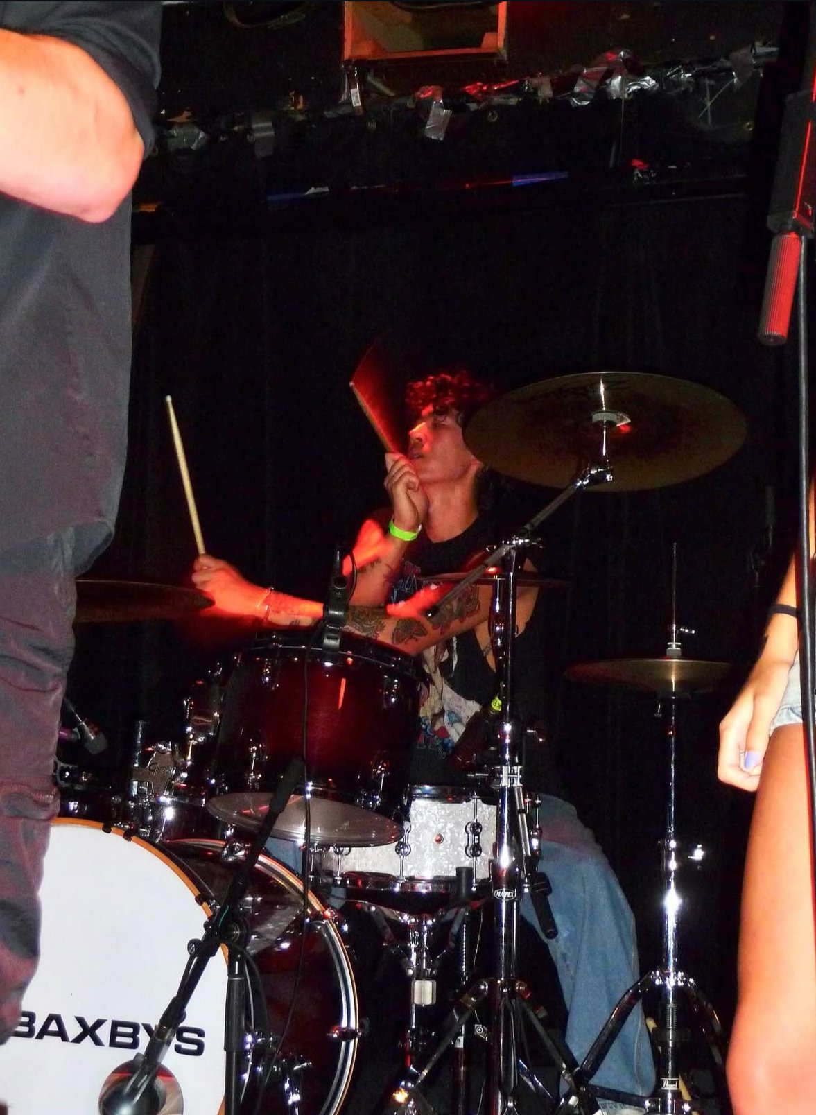 A person with tattoos and curly hair playing a drum set on stage, holding a red object near their face, with dark background and other individuals partially visible nearby.