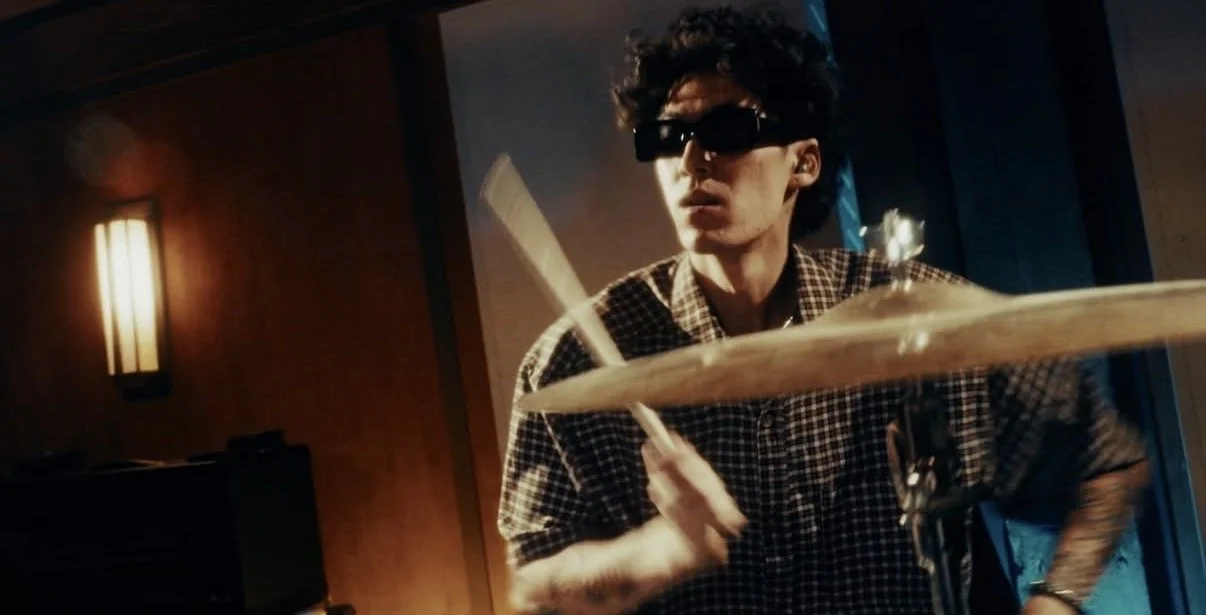 A young man with dark curly hair wearing sunglasses and a checkered shirt, playing the drums in a dimly lit room.