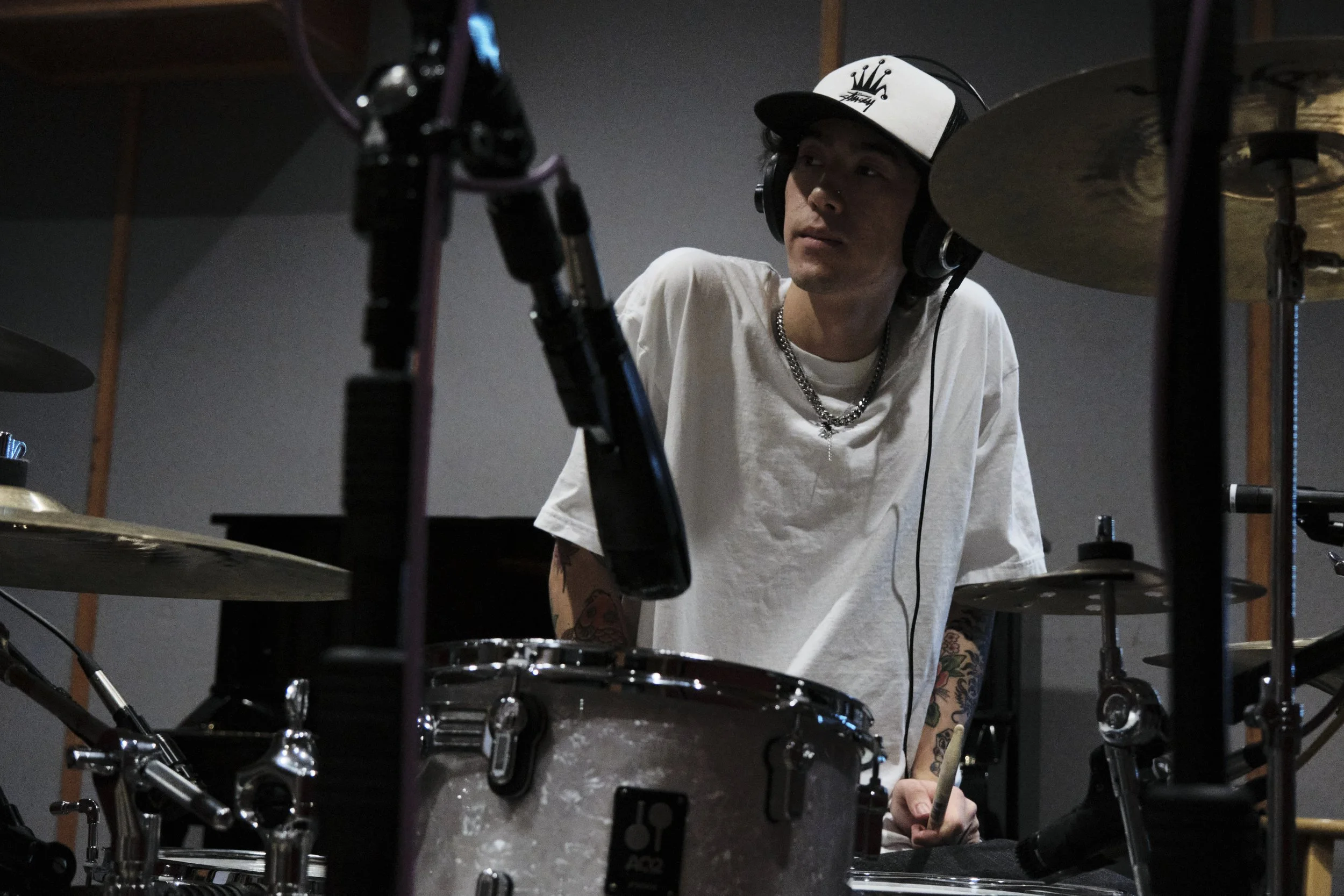 Young man with tattoos and a baseball cap playing drums in a dimly lit studio.